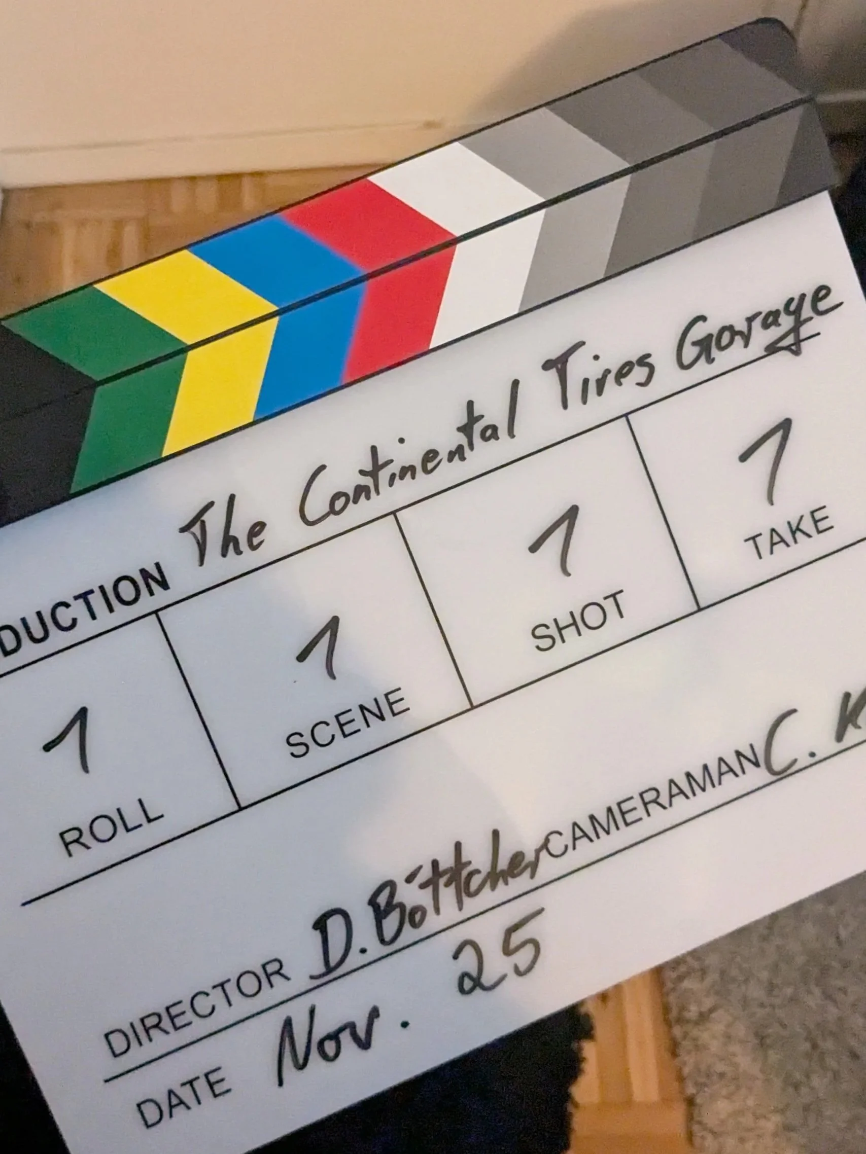 Clapboard with colored stripes and handwritten text for a film production, titled 'The Continental Tire Garage.' It includes details such as roll, scene, shot, take, director, and date, with the director's name 'D. Bottcher' and date 'Nov. 25'.