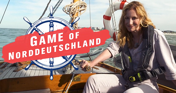 A woman sitting on a sailboat during daytime, with the sea and a distant shoreline in the background. Overlay text reads 'Game of Norddeutschland' next to a nautical wheel graphic.