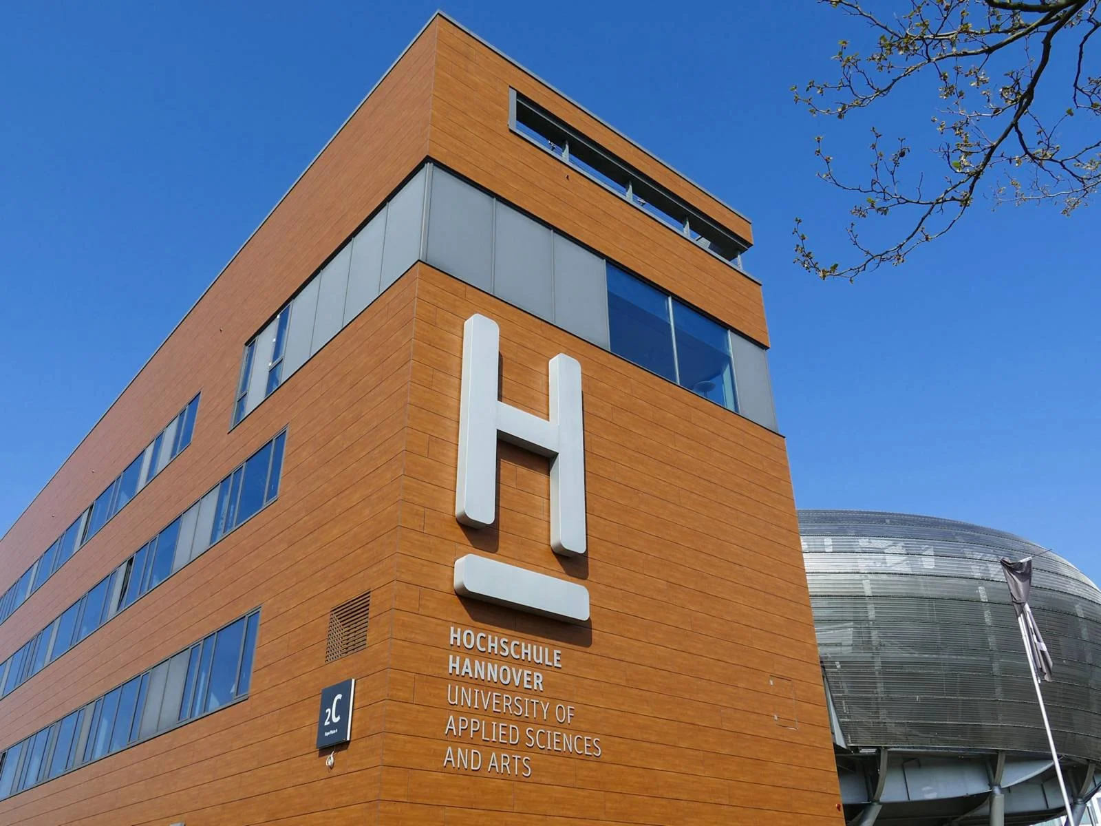 Modern university building with a large white 'H' sign on orange/brown exterior wall. Sign reads 'Hochschule Hannover University of Applied Sciences and Arts.' Blue sky and a part of a tree are visible.