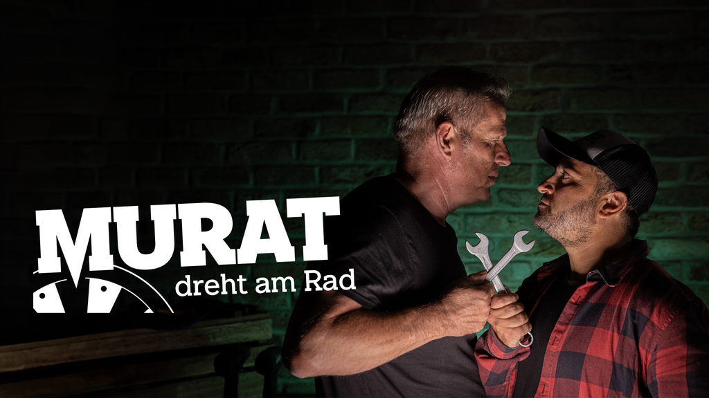 Two men face each other, one older and one younger, holding wrenches. The older man has gray hair and is wearing a black shirt, while the younger man has a beard, is wearing a cap and a red plaid shirt. They are standing in front of a dark brick wall, with a logo and text that says "Murat dreht am Rad."