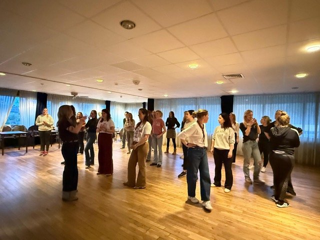 Group of women participating in a dance class or social dance event in a spacious room with wooden floors and large windows.