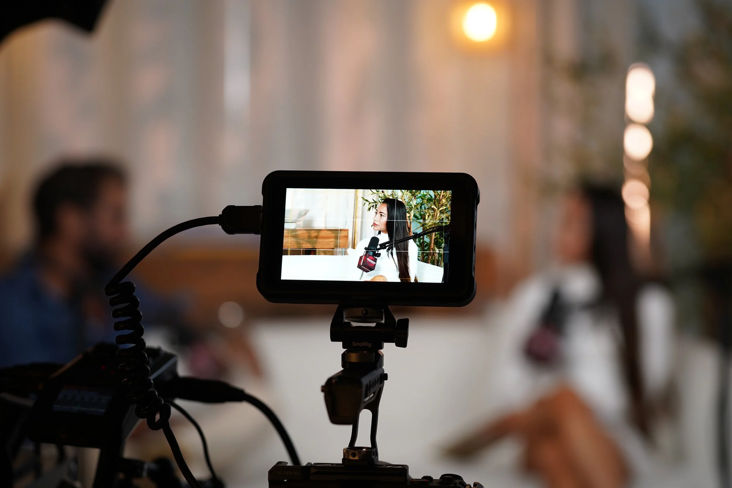 A woman with long black hair is being filmed by a camera with a microphone attached, seen on a camera screen showing her face in focus, in an indoor setting with a plant background.