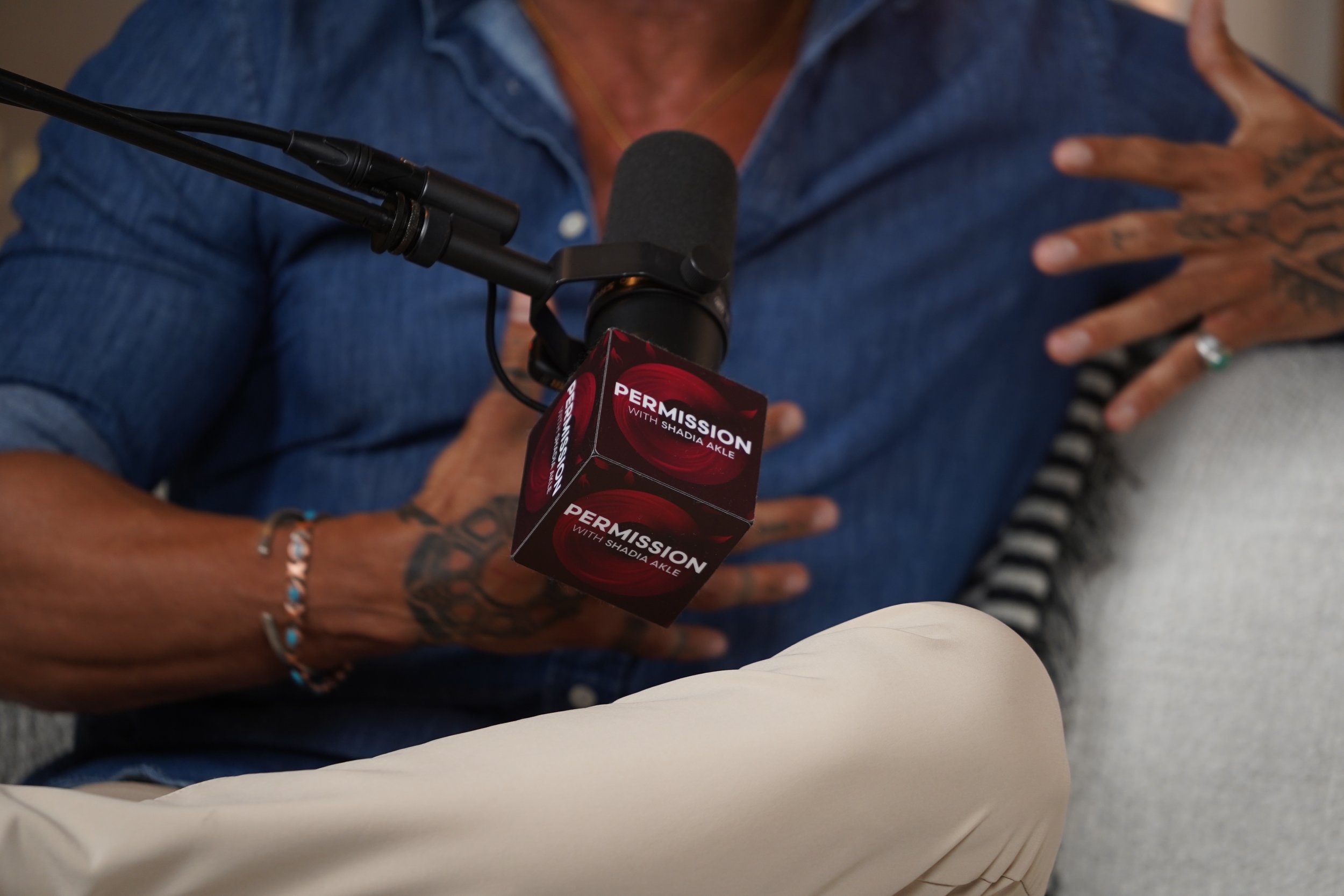 A person wearing a blue denim shirt is speaking into a microphone labeled 'Permission with Shadia Akle.' The person has tattoos on their hand and wrist, and is wearing rings and bracelets. A person in a beige jacket is sitting nearby.