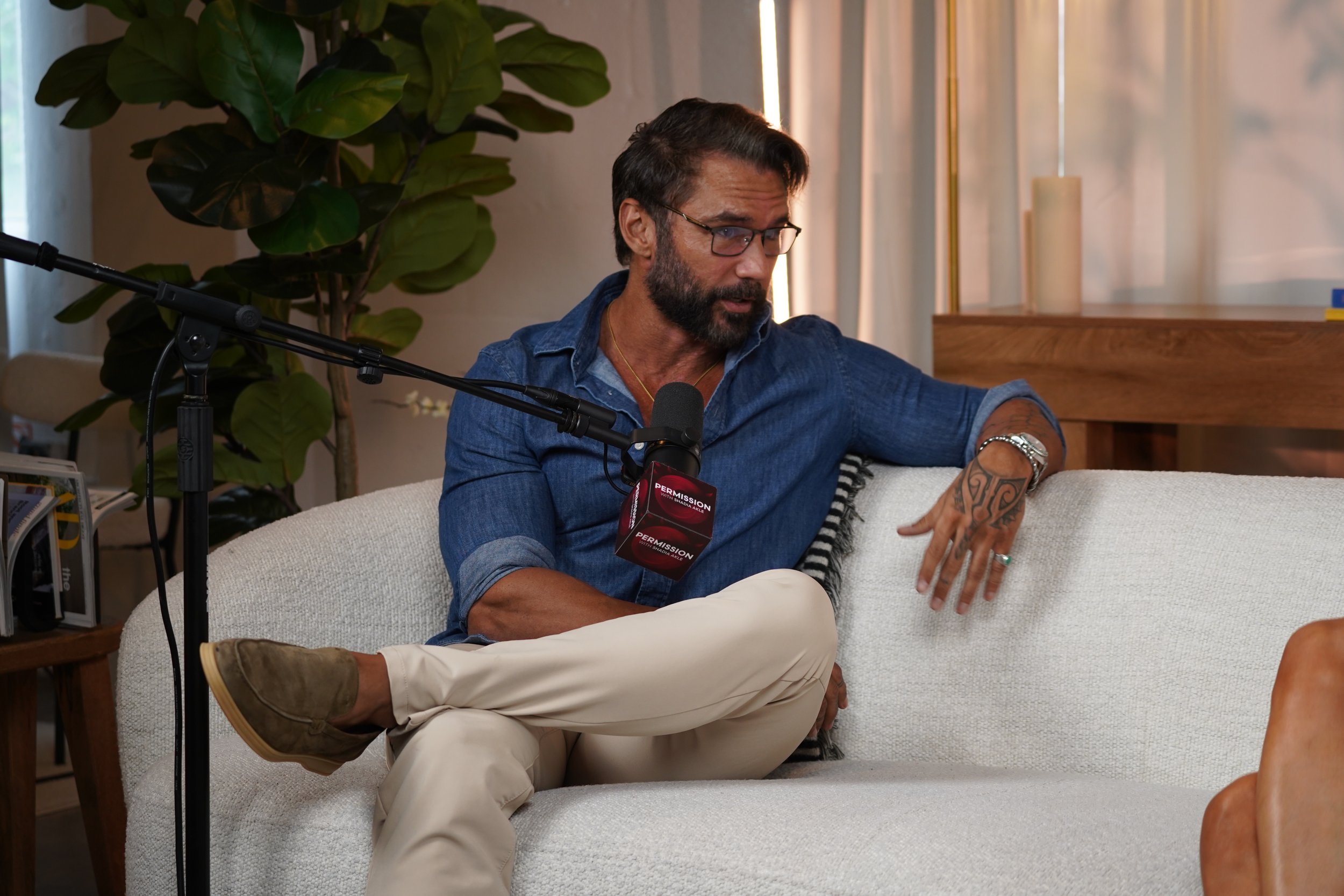A man with dark hair, glasses, and a beard sitting on a white sofa, with a green plant and a wooden table in the background. He is wearing a blue shirt and beige pants, and appears to be involved in a conversation or interview with recording equipmen