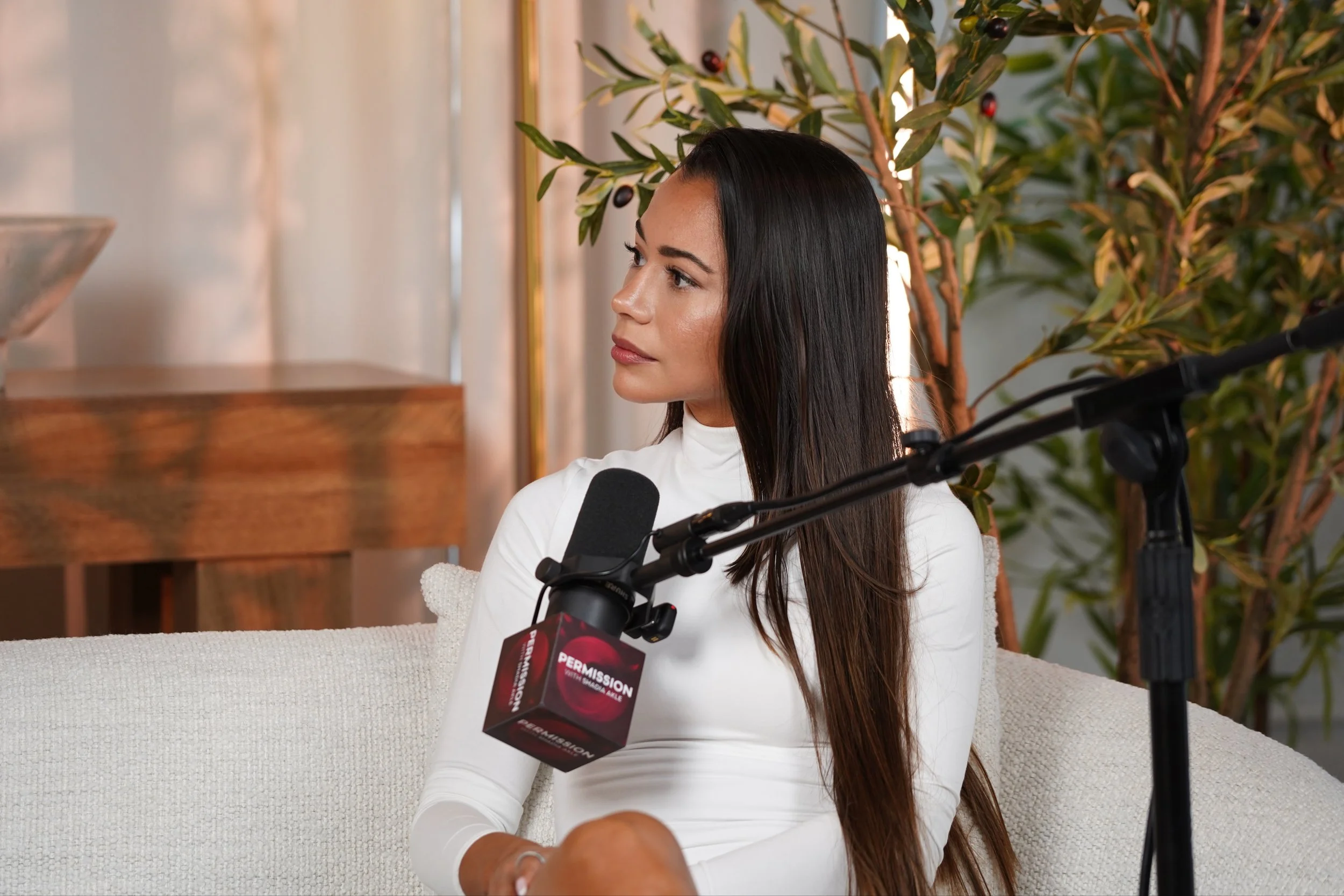 A woman with long dark hair and light skin sitting on a beige couch, wearing a white long-sleeve top, and speaking into a black microphone with a red and black label that reads 'PERMISSION.' She is in a well-lit room with green leafy plants and a woo