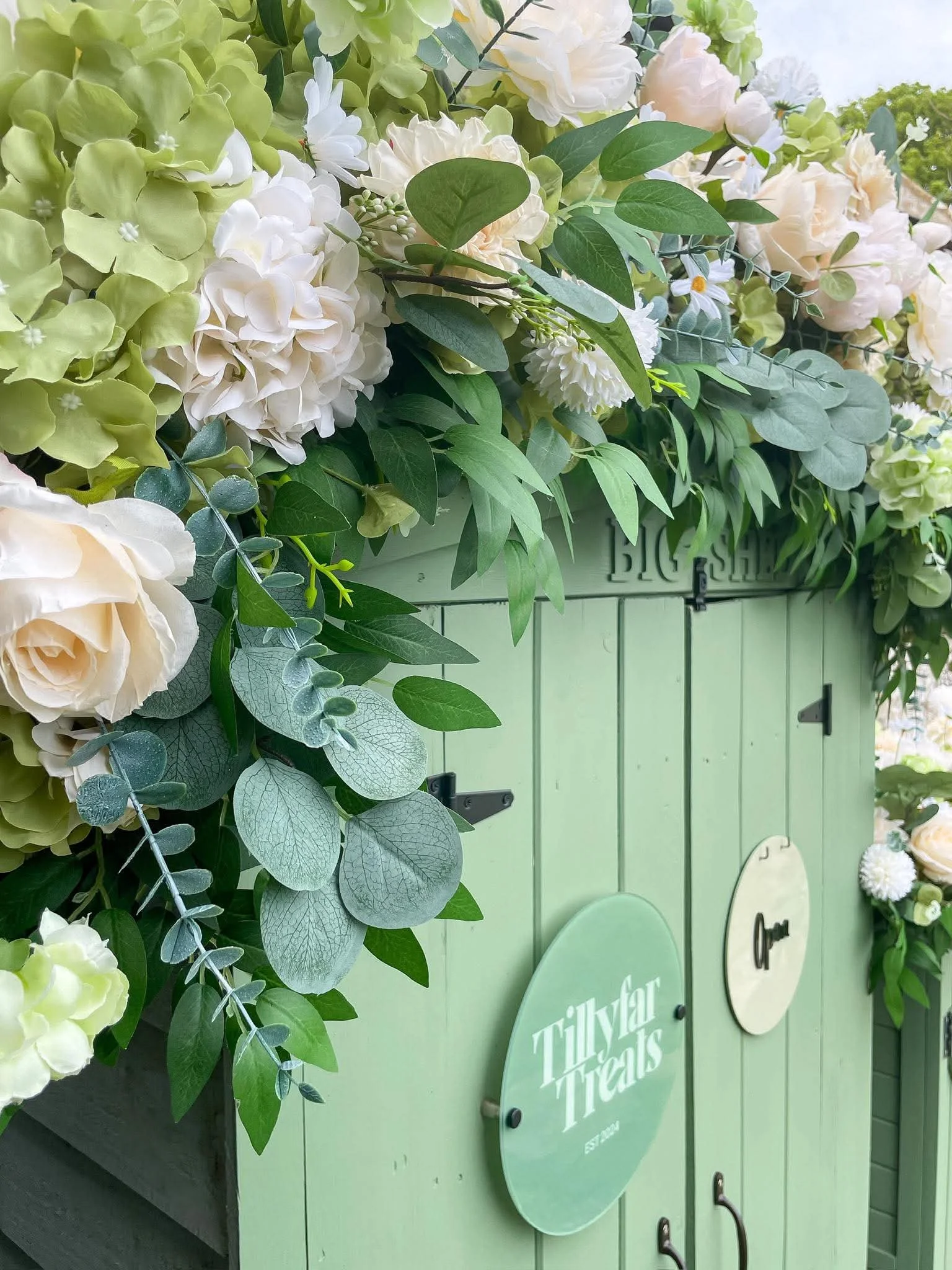 A floral arrangement of white roses, green hydrangeas, and assorted green leaves, displayed against a pastel green wooden backdrop with signs.
