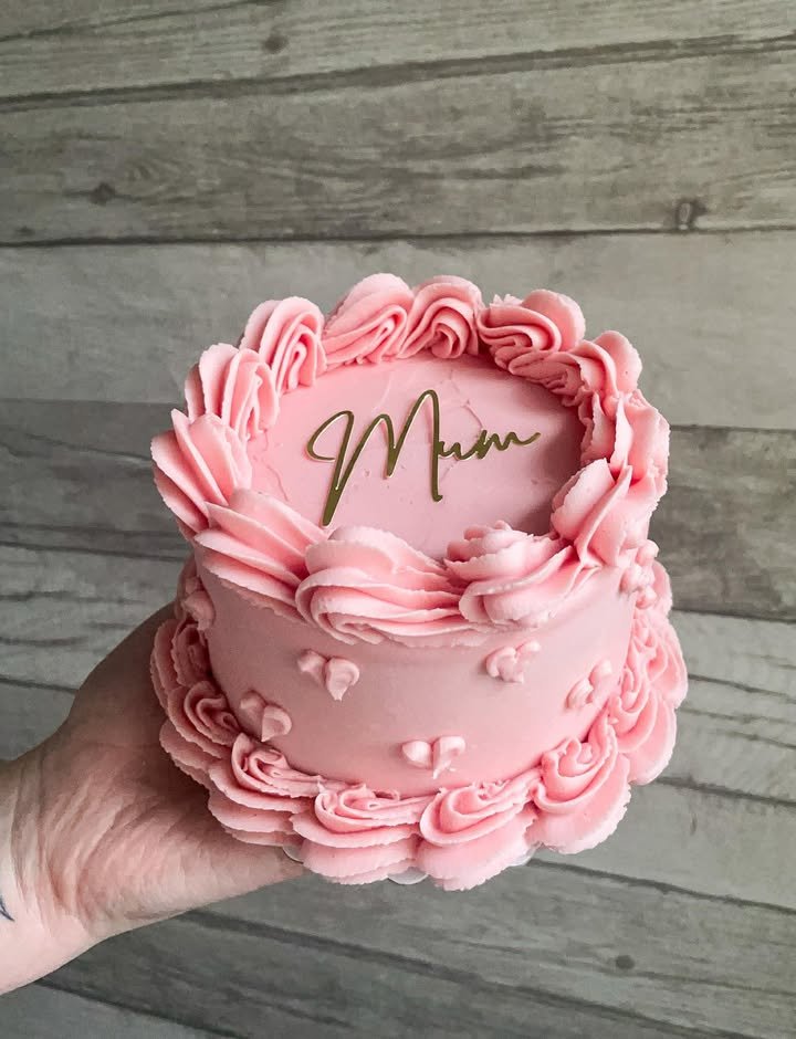Pink cake with decorative frosting and the word 'Mum' written on top, held in a person's hand against a wooden background.