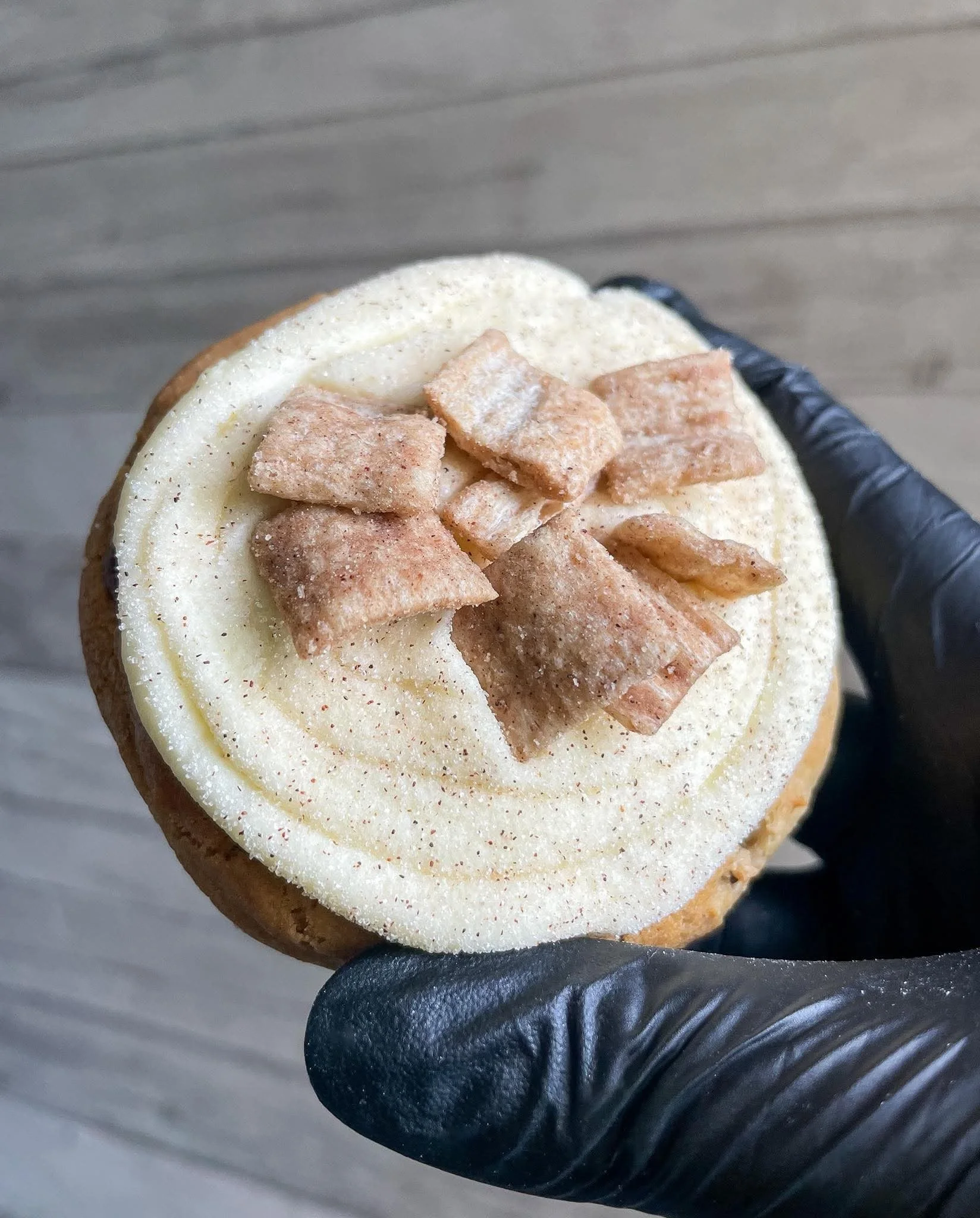 Close-up of a person's hand wearing a black glove holding a sliced bagel topped with cream cheese and small square cereal pieces.