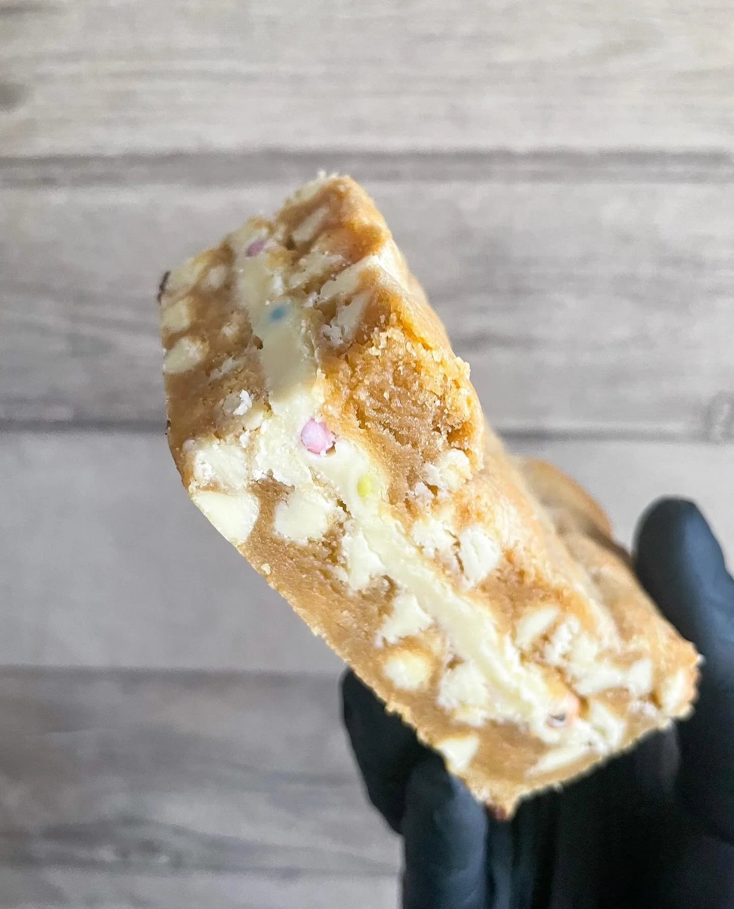Close-up of a partially eaten ice cream bar with a crunchy caramelized coating and visible bits of nuts on top, being held by a person wearing a black glove against a gray background.