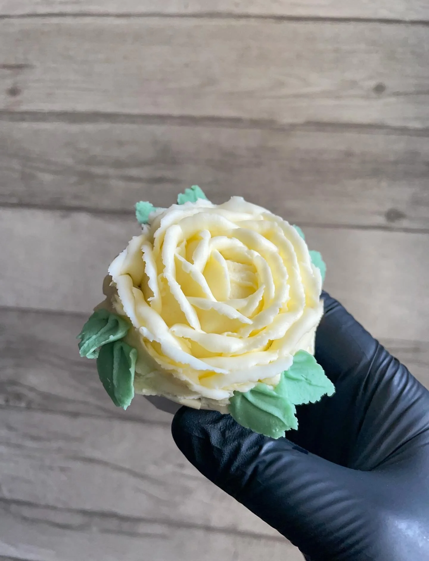 Hand holding a cupcake decorated with a yellow rose made from frosting and green frosting leaves, against a wooden background.