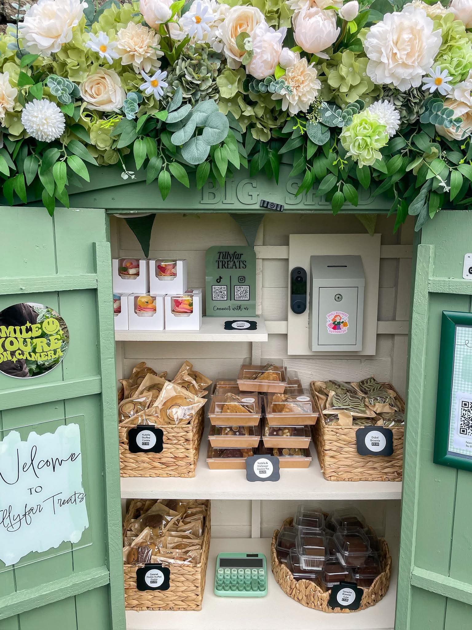 A display of various sweet treats in small boxes and containers, including caramel stuffed cookies, Nutella and marshmallow cookies, ganache brownies, and other assorted baked goods, arranged on shelves and in baskets with a decorative sign above.