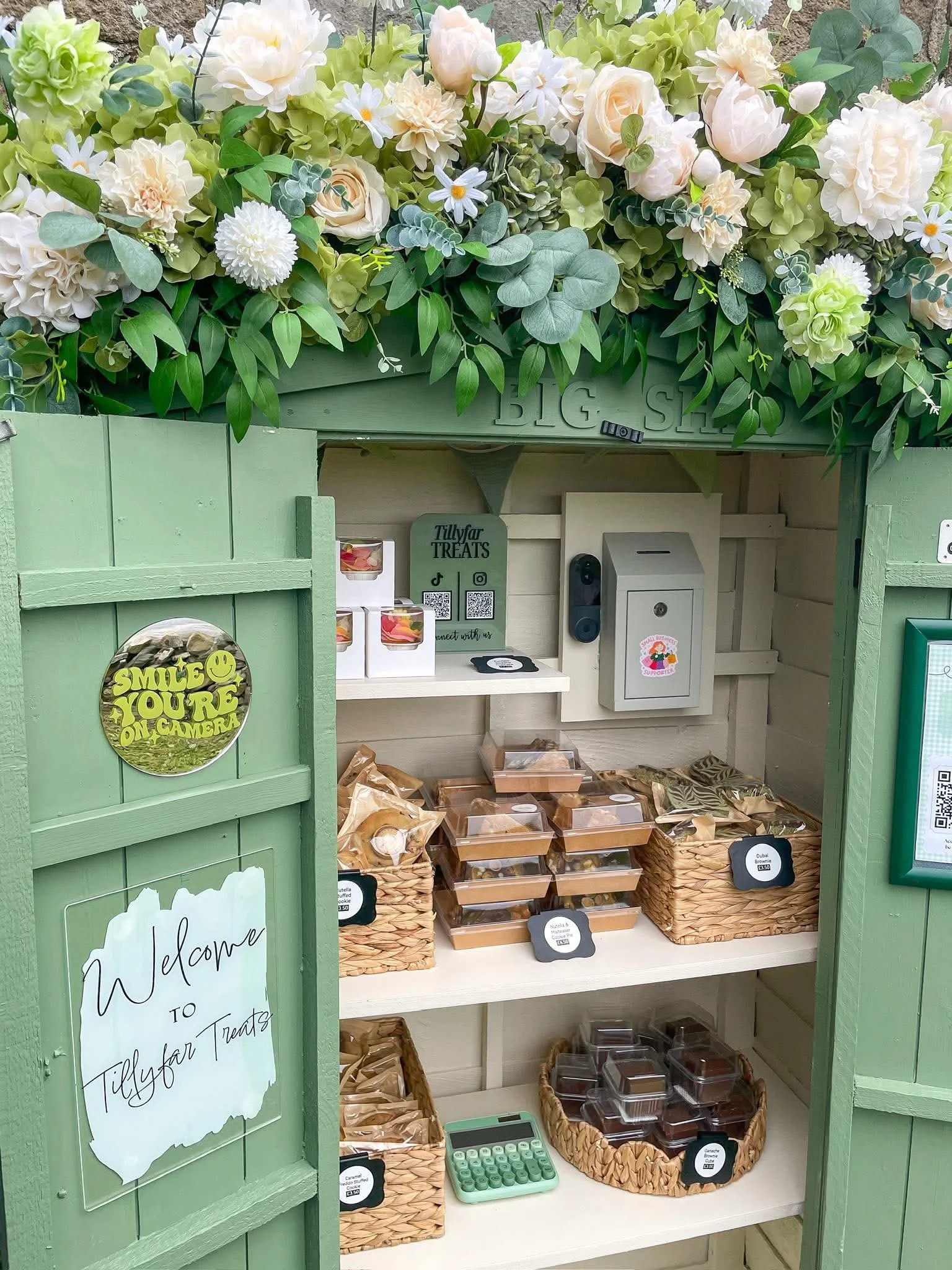 Display of packaged cookies and treats in a small shop, with a floral arrangement on top, a sign that says 'Welcome to Tillyfar Treats,' and baskets of assorted cookies and desserts.
