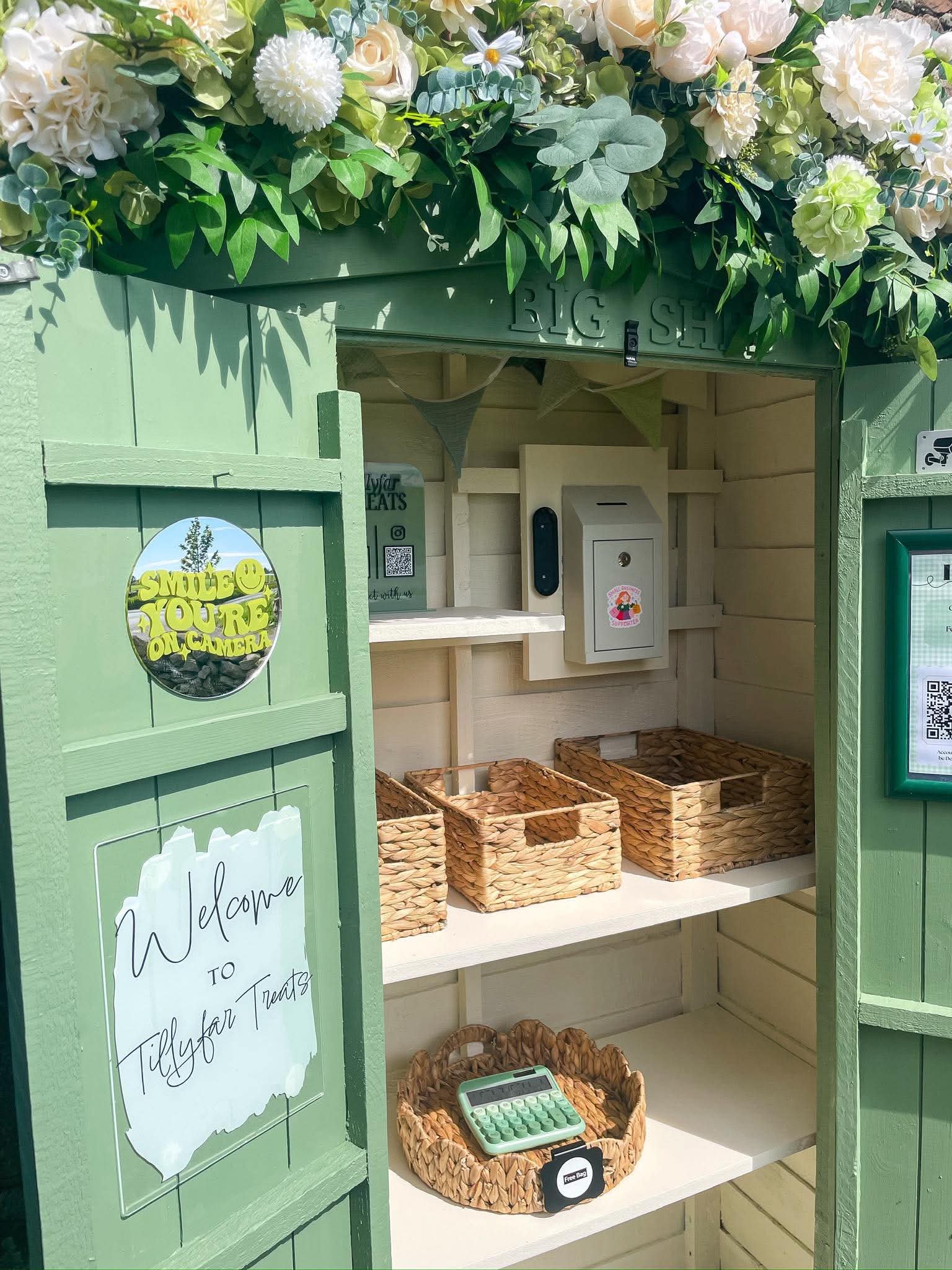 Small green outdoor stand with a floral topping, open shelves with empty woven baskets, a small calculator, and signs welcoming visitors.