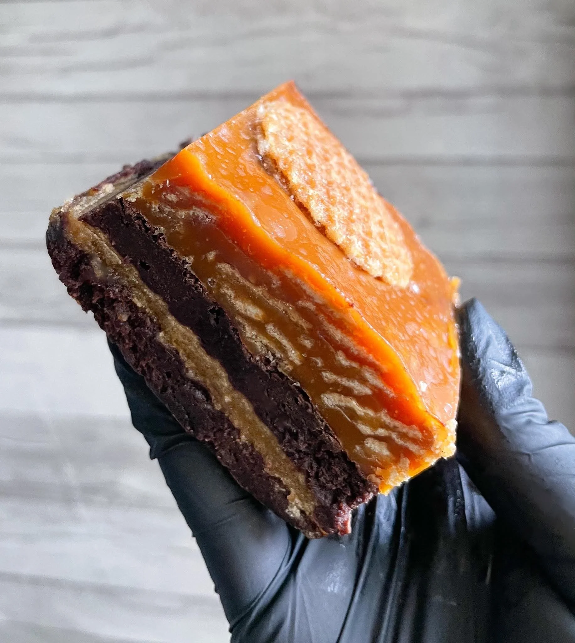 Close-up of a piece of layered dessert with a dark brownie bottom, chocolate filling, caramel layer, and topping, being held by a person wearing a gray glove.