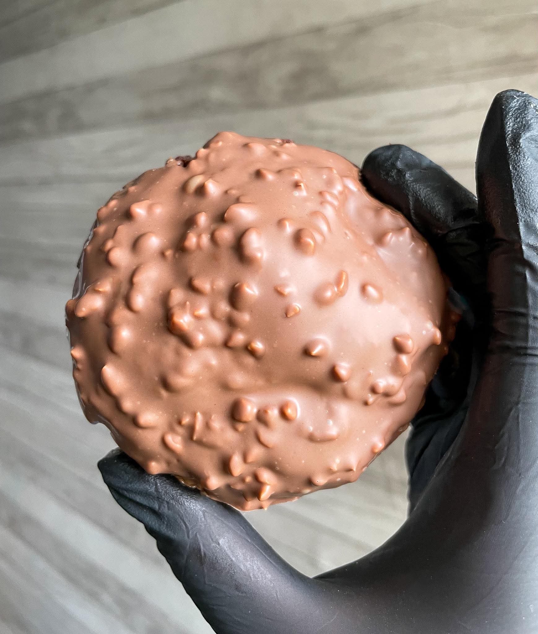 Close-up of a round chocolate-covered confection with a bumpy textured surface, being held with black gloves, against a gray wooden background.