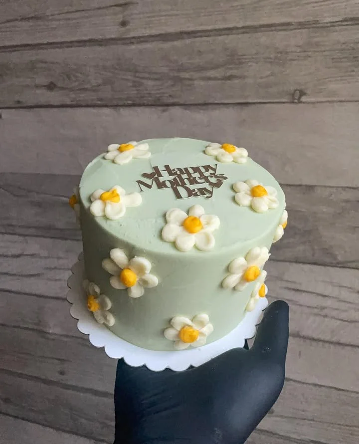 A round cake with pale green frosting decorated with white daisies with yellow centers, topped with a "Happy Mother’s Day" chocolate message, being held by a person wearing black gloves, against a wooden background.
