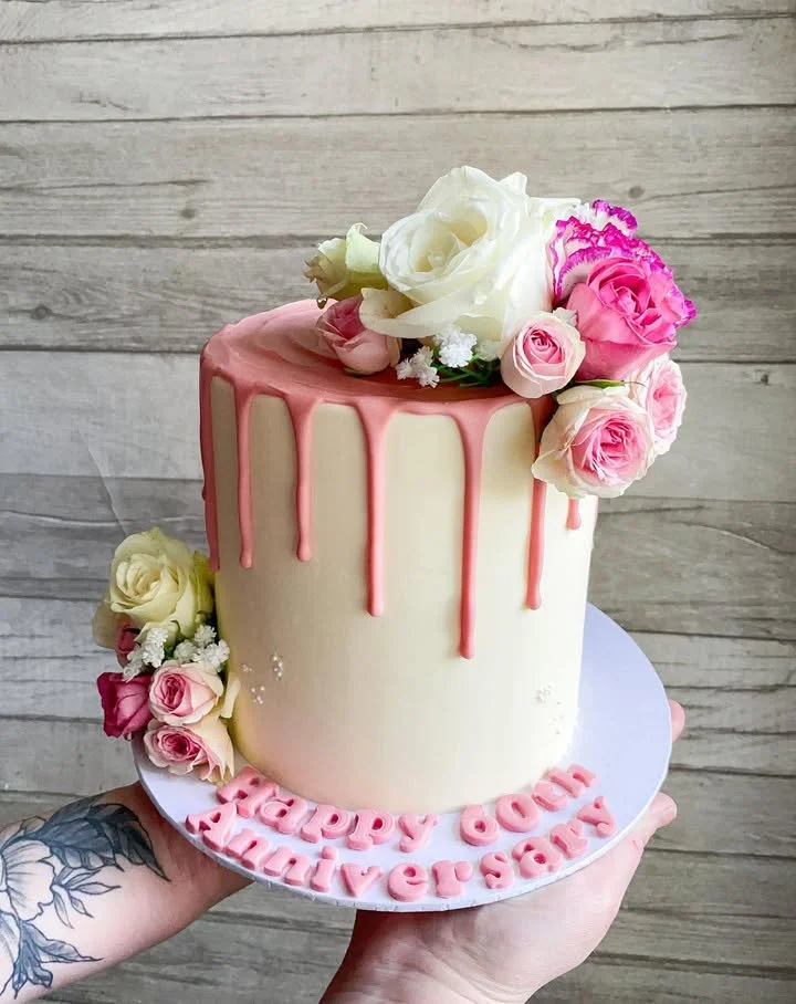 A decorated birthday cake with white and pink frosting, surrounded by pink and white flowers, and pink lettering that reads "HAPPY GOM SURIYA".