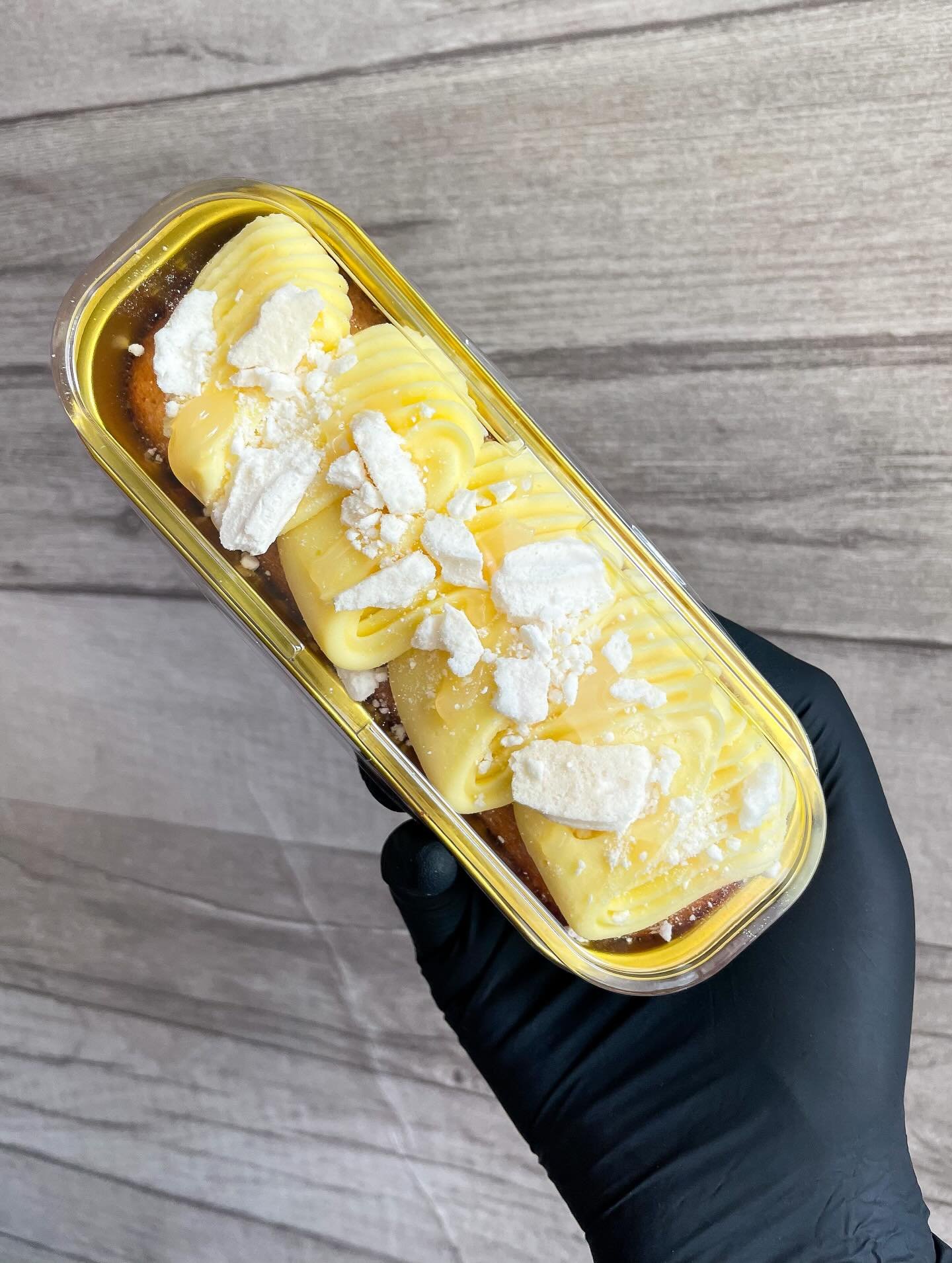 Hand holding a plastic container with yellow topped dessert, garnished with white crumbles, against a wooden background.