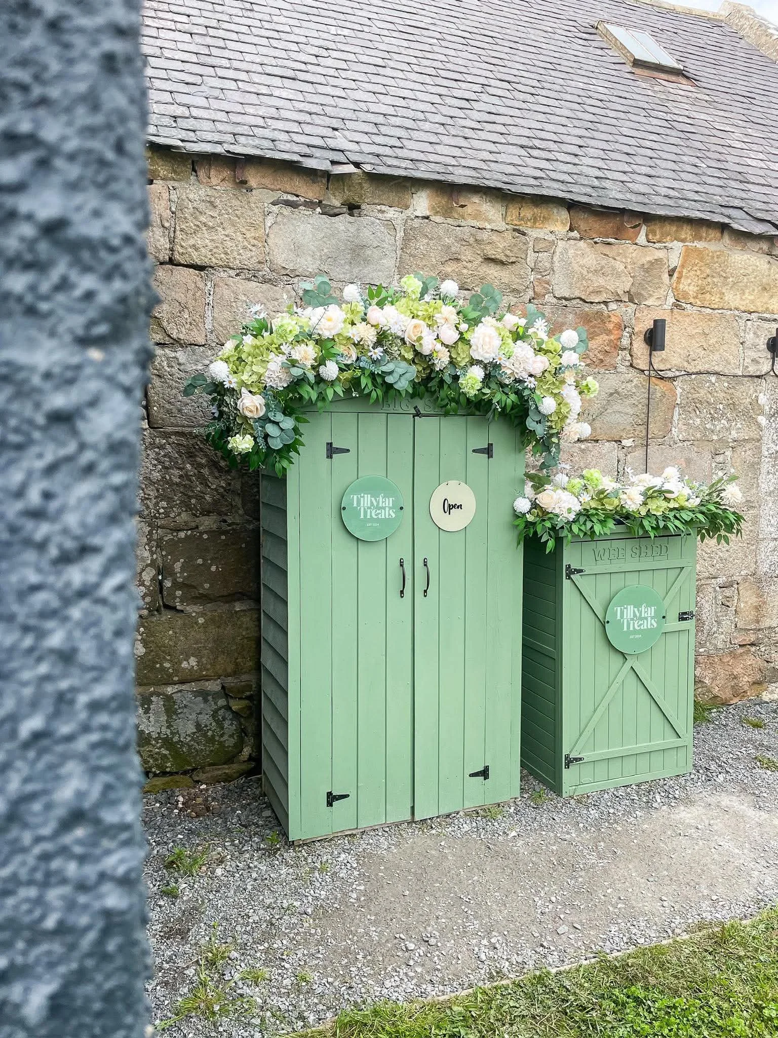 Two green wooden storage sheds with floral decorations on top, labeled 'Tillyar Treats' and 'WEE SHED,' set against a stone wall with a small skylight in the roof.