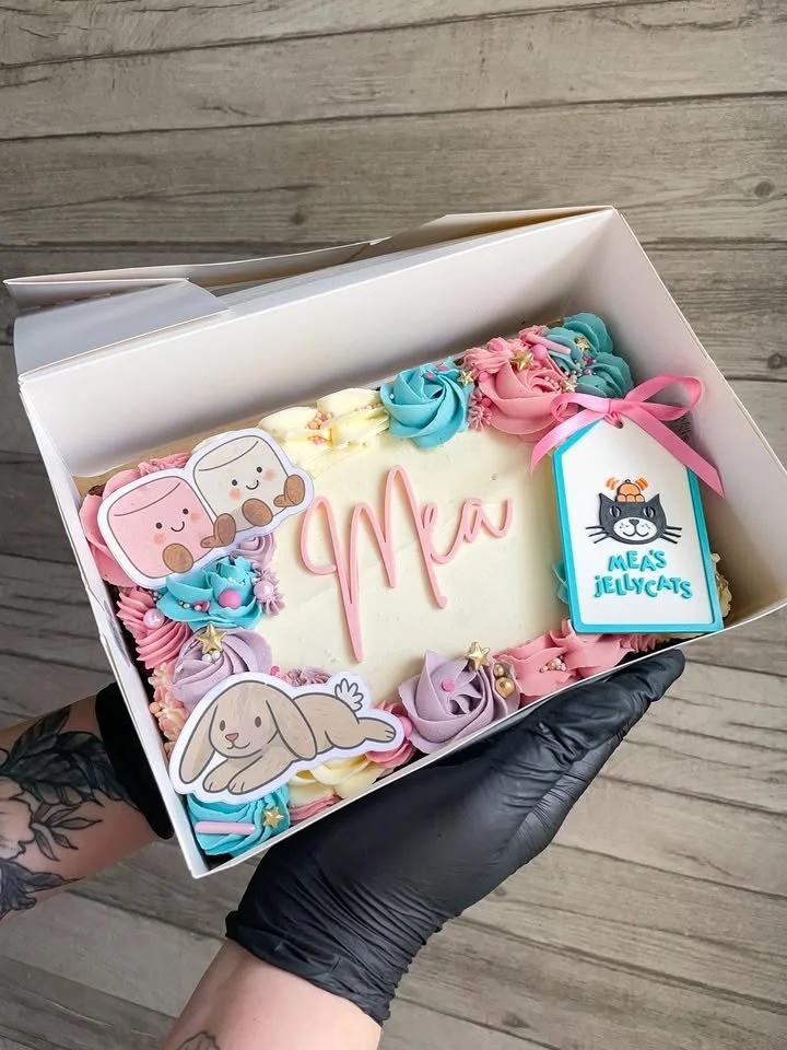 Decorated birthday cake with pink, blue, and purple frosting, topped with cartoon dog and cat toppers, with the name 'Mia' written in pink icing, inside a white box on a wooden floor.