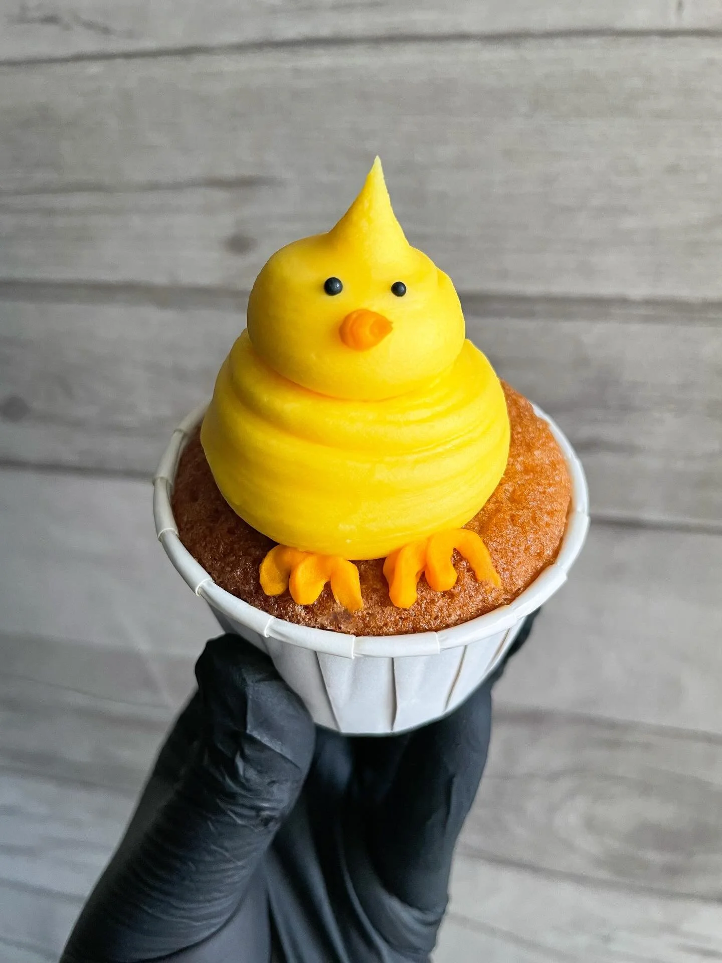 Ice cream cone decorated to look like a yellow chick with black eyes, an orange beak, and small orange feet, set on top of a baked good.