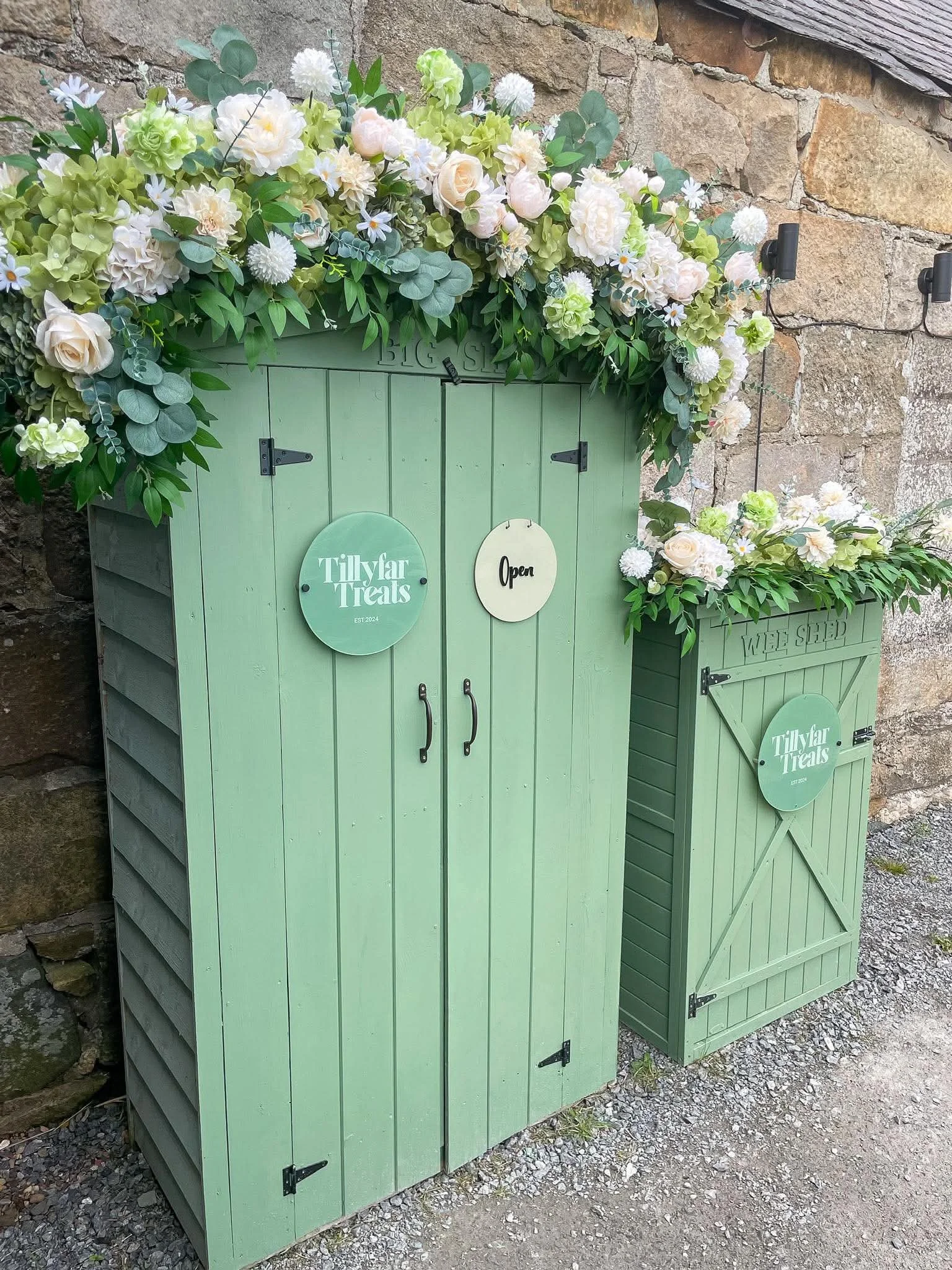 Green wooden cake sheds with flower arrangements on top, placed outdoors against a stone wall.