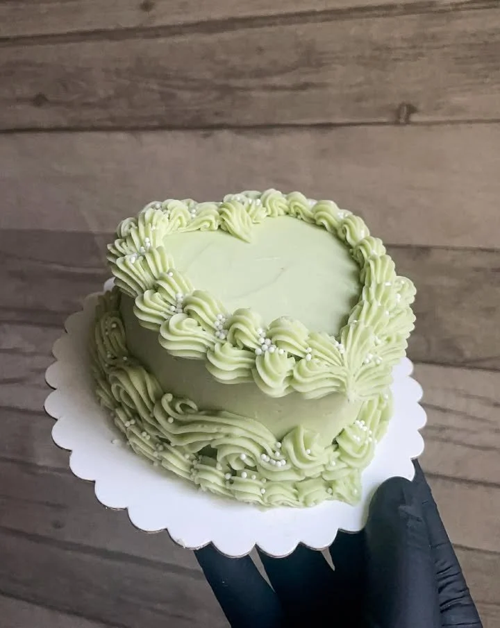 A heart-shaped cake decorated with pale green piping and topped with silver pearl sprinkles, held on a white scalloped cake board.
