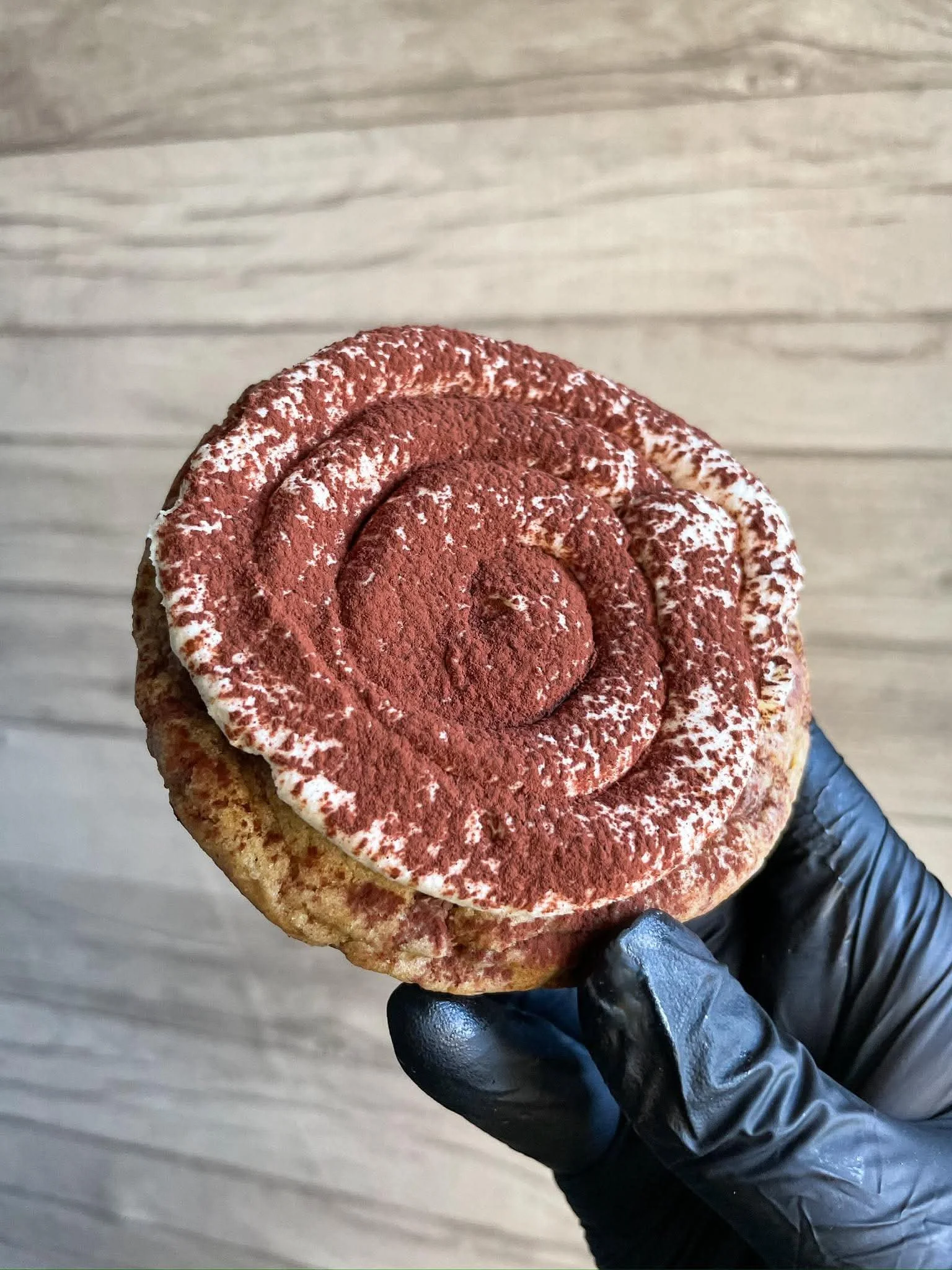 A person wearing a black glove holding a tiramisu dessert in a clear cup with a wooden surface background. The dessert has layers of cream, cocoa powder, and sponge cake.