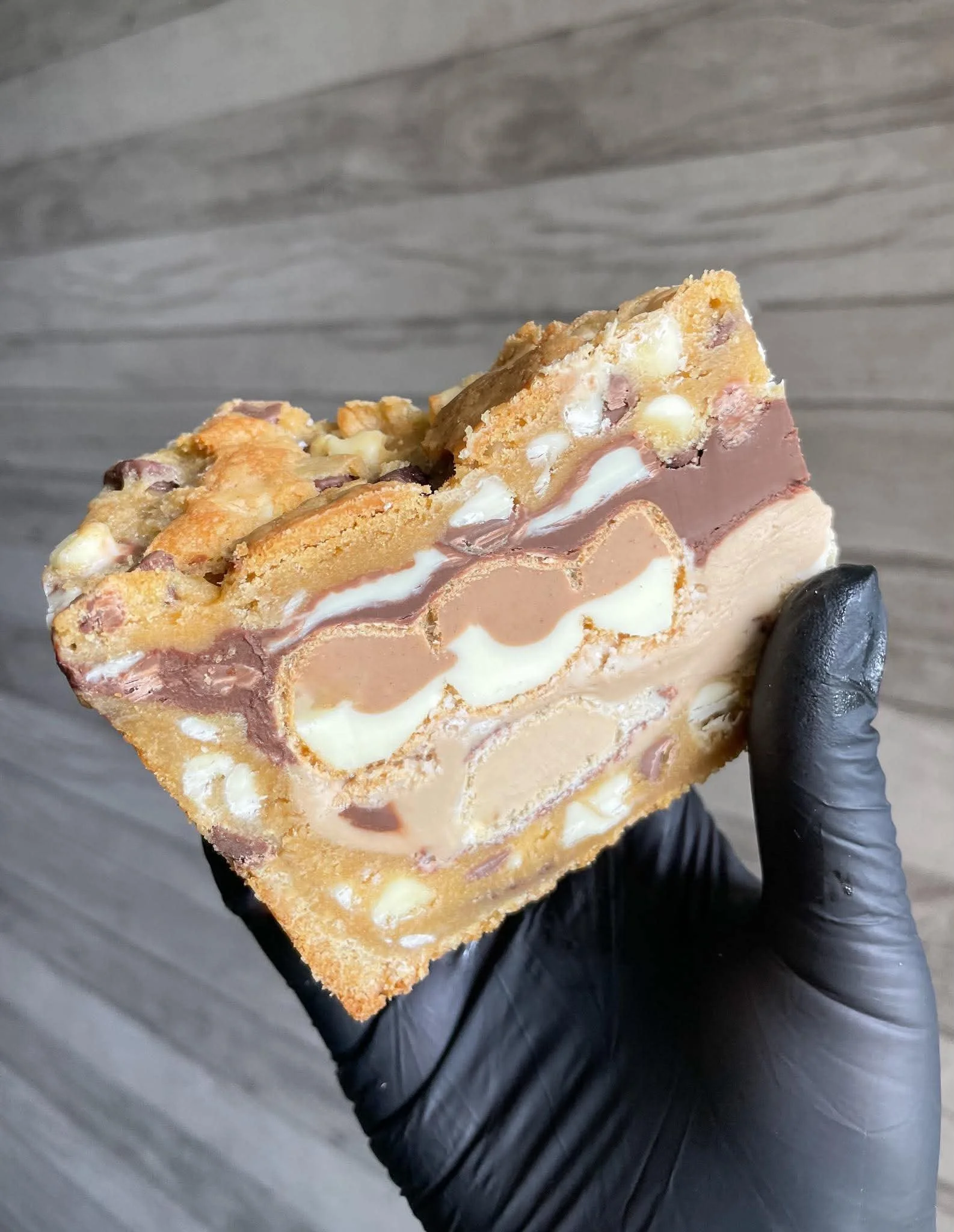 A hand wearing a black glove holds a slice of layered dessert with nuts, chocolate, and cream layers on a gray surface background.