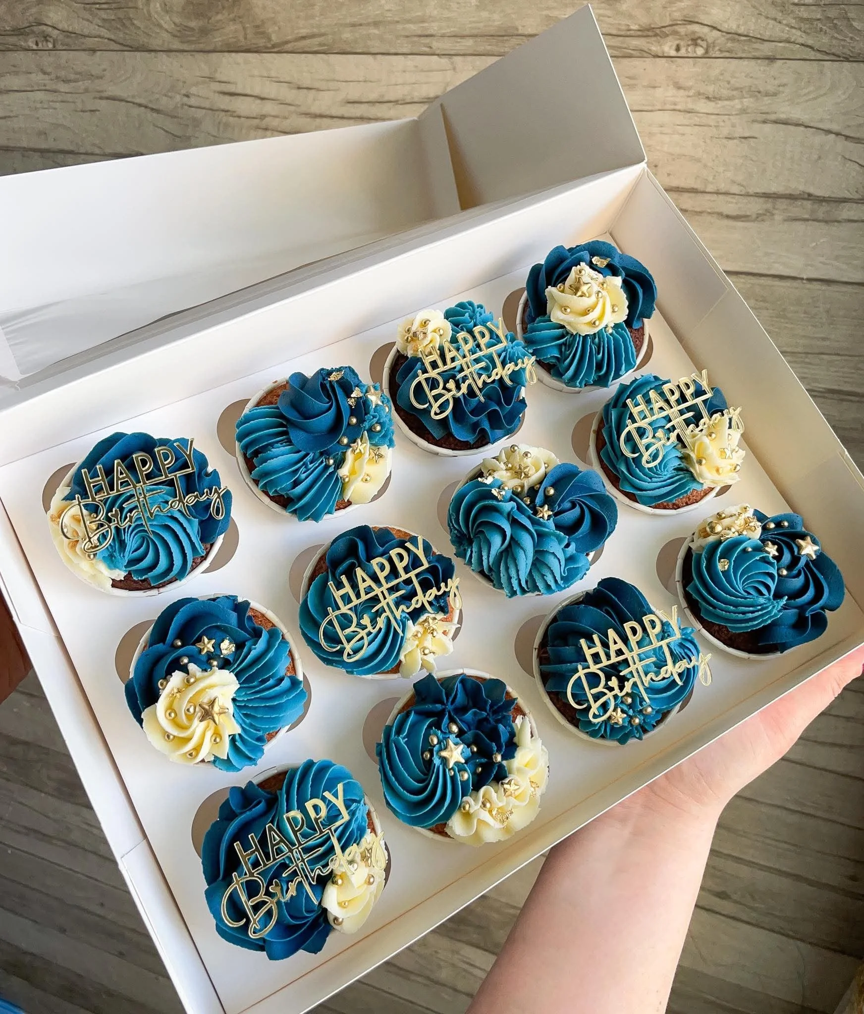 A box of decorated cupcakes with blue and white frosting, adorned with gold and sprinkles, including "Happy Birthday" signs for celebration.