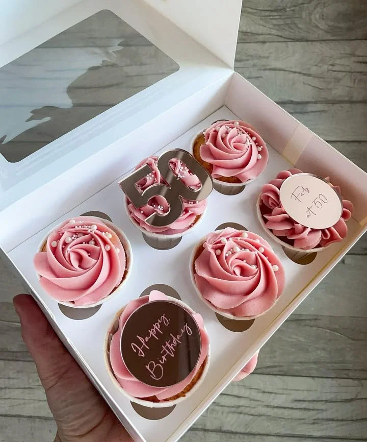 Six birthday cupcakes in a white box featuring pink frosting decorated with small white sprinkles, with one cupcake topped with a metallic '30' cake topper and another with a round chocolate disc that says 'Happy Birthday'; two cupcakes have round wh