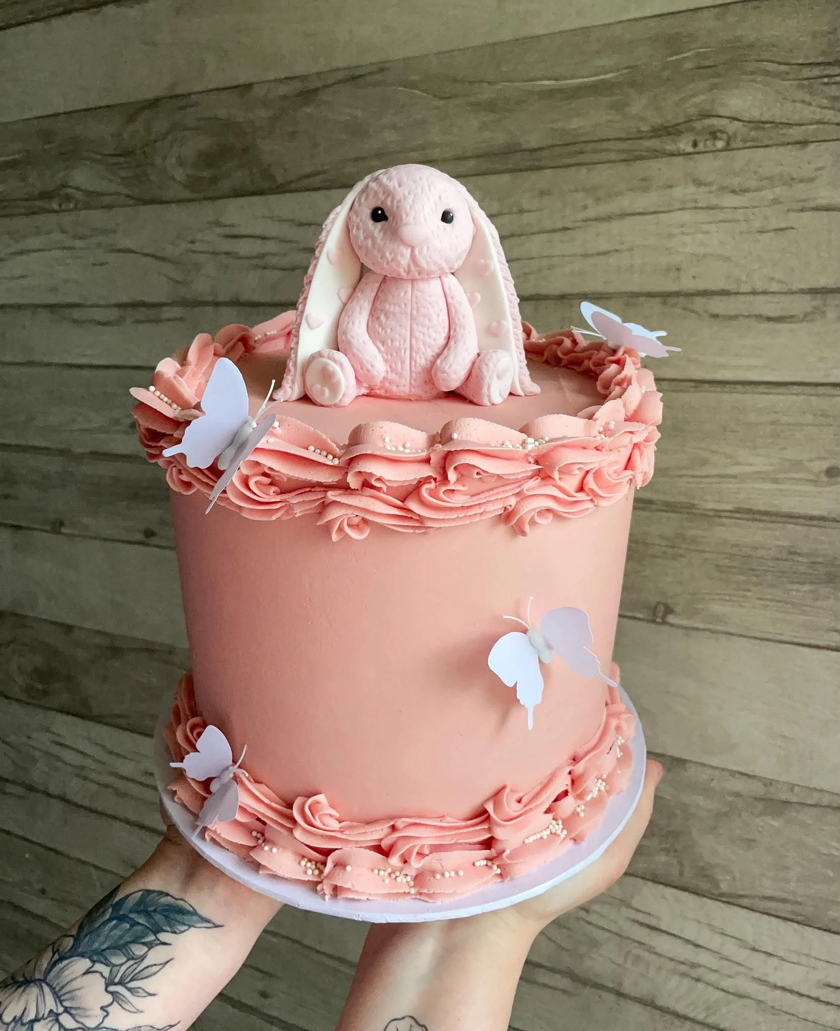 Pink layered cake decorated with white butterflies, featuring a central bunny figure on top, set against a wooden background.