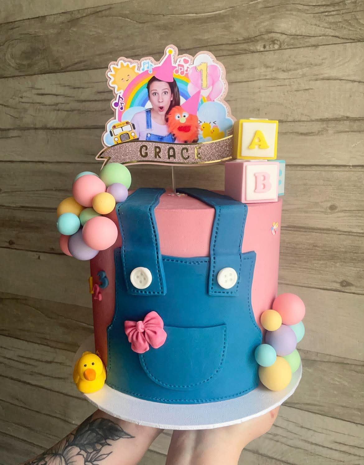 A colorful cake designed for a child's birthday celebration. The cake has a pink base with a blue fondant pocket and straps, resembling overalls. It is decorated with pastel-colored ball embellishments, a small rubber duck, and a pink bow. A topper f