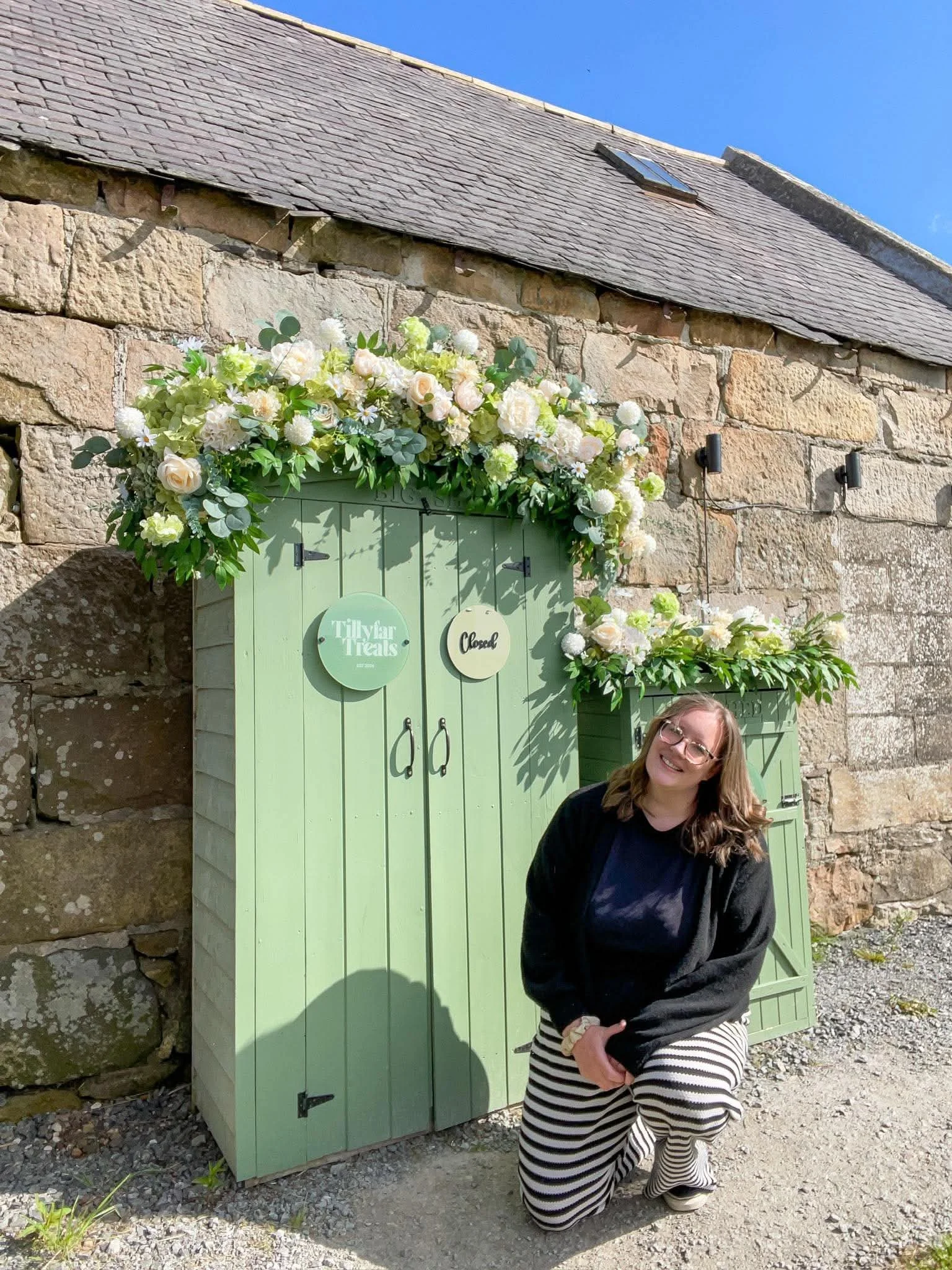 A woman kneeling and smiling in front of a light green outdoor cake shed with floral decorations, against a stone building wall under a clear blue sky.