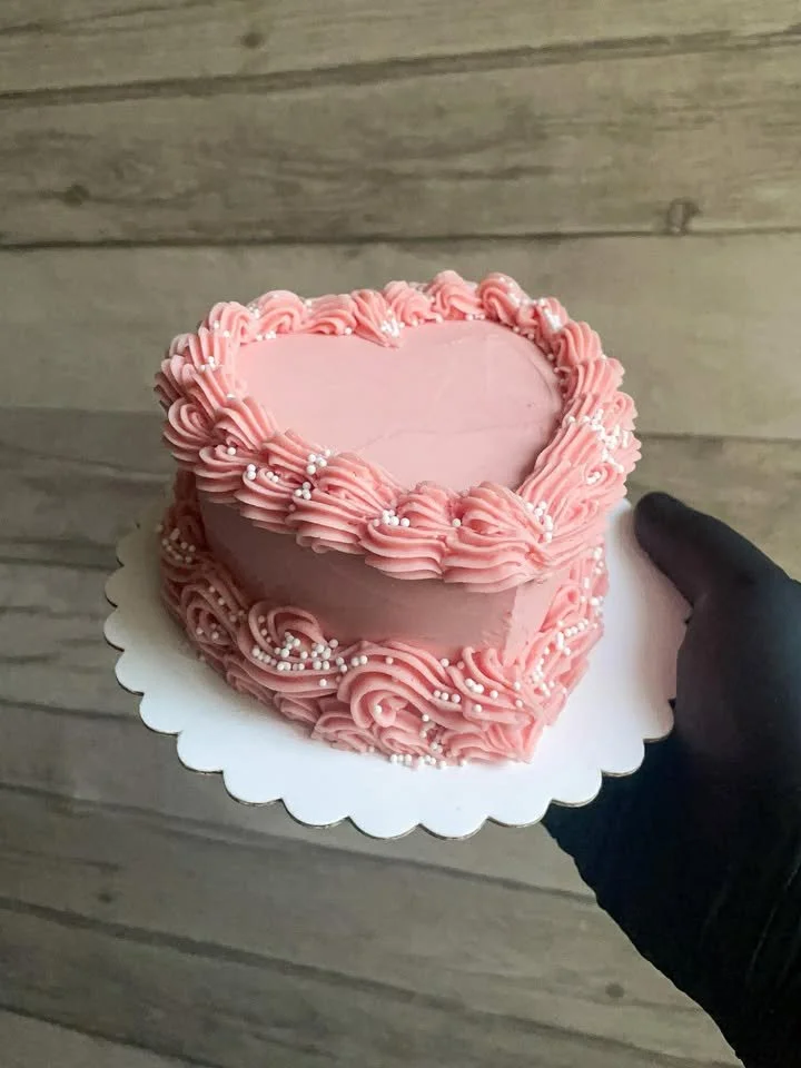 A person holding a heart-shaped pink frosted cake decorated with piped pink icing and small white pearls, standing on a wooden floor.