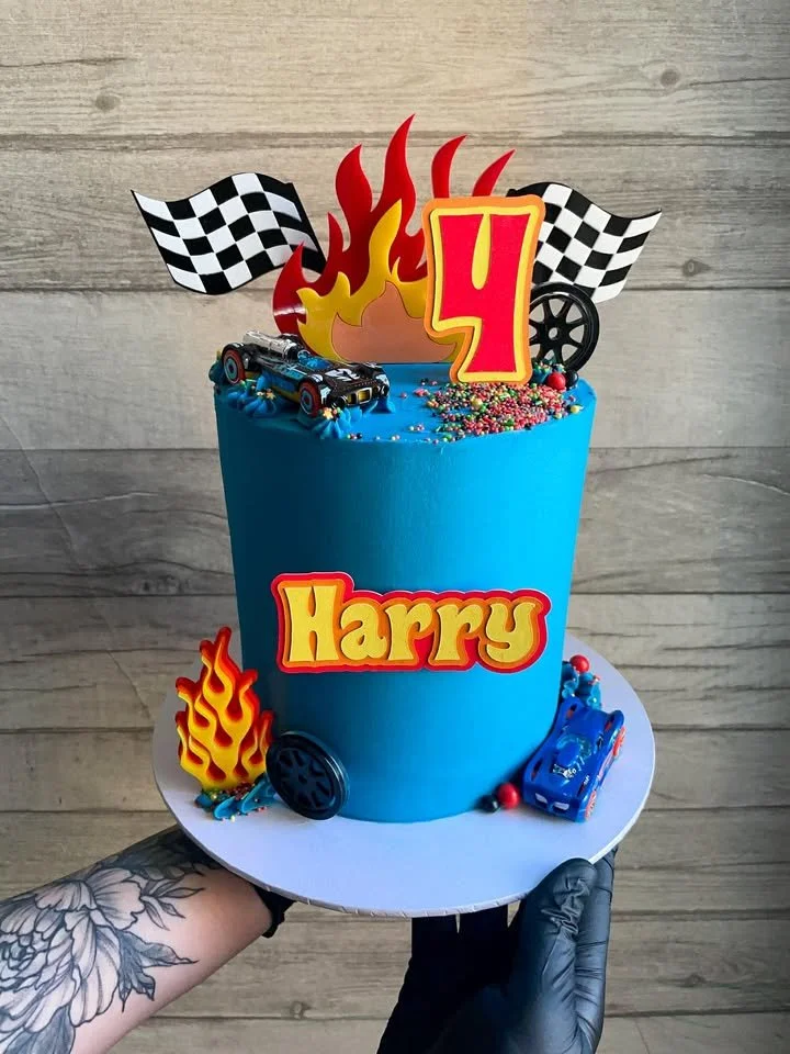 Blue birthday cake with racing-themed decorations, including checkered flags, flames, a number 4, a toy race car, and vibrant sprinkles, held by a person wearing black gloves.