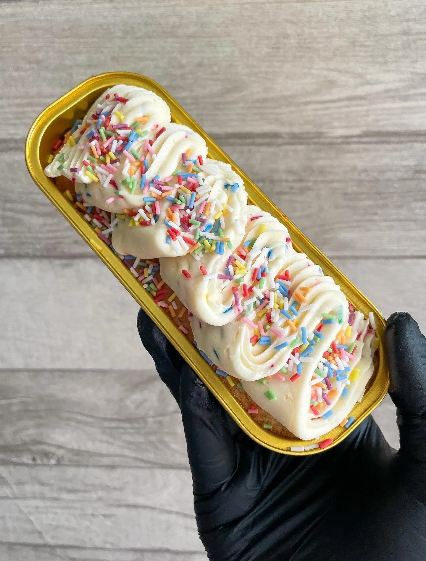 A hand wearing a black glove holding a gold-colored phone case with a dessert inside, featuring piped white frosting and rainbow sprinkles, on a gray wooden surface.