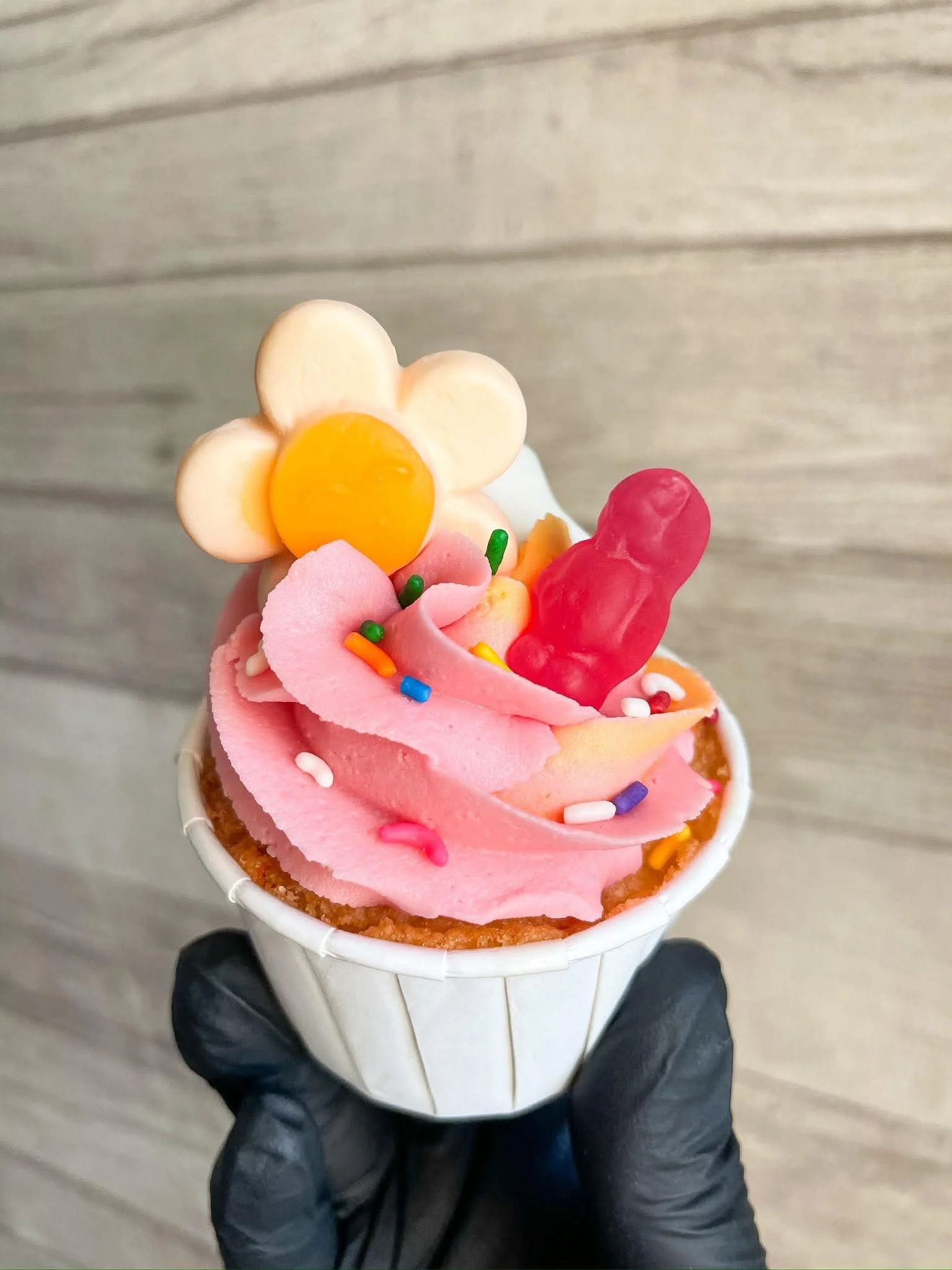 A person wearing black gloves holds a cup of pink ice cream with colorful sprinkles, topped with a flower-shaped cookie, a yellow candy, and a red gummy candy, against a wood floor background.