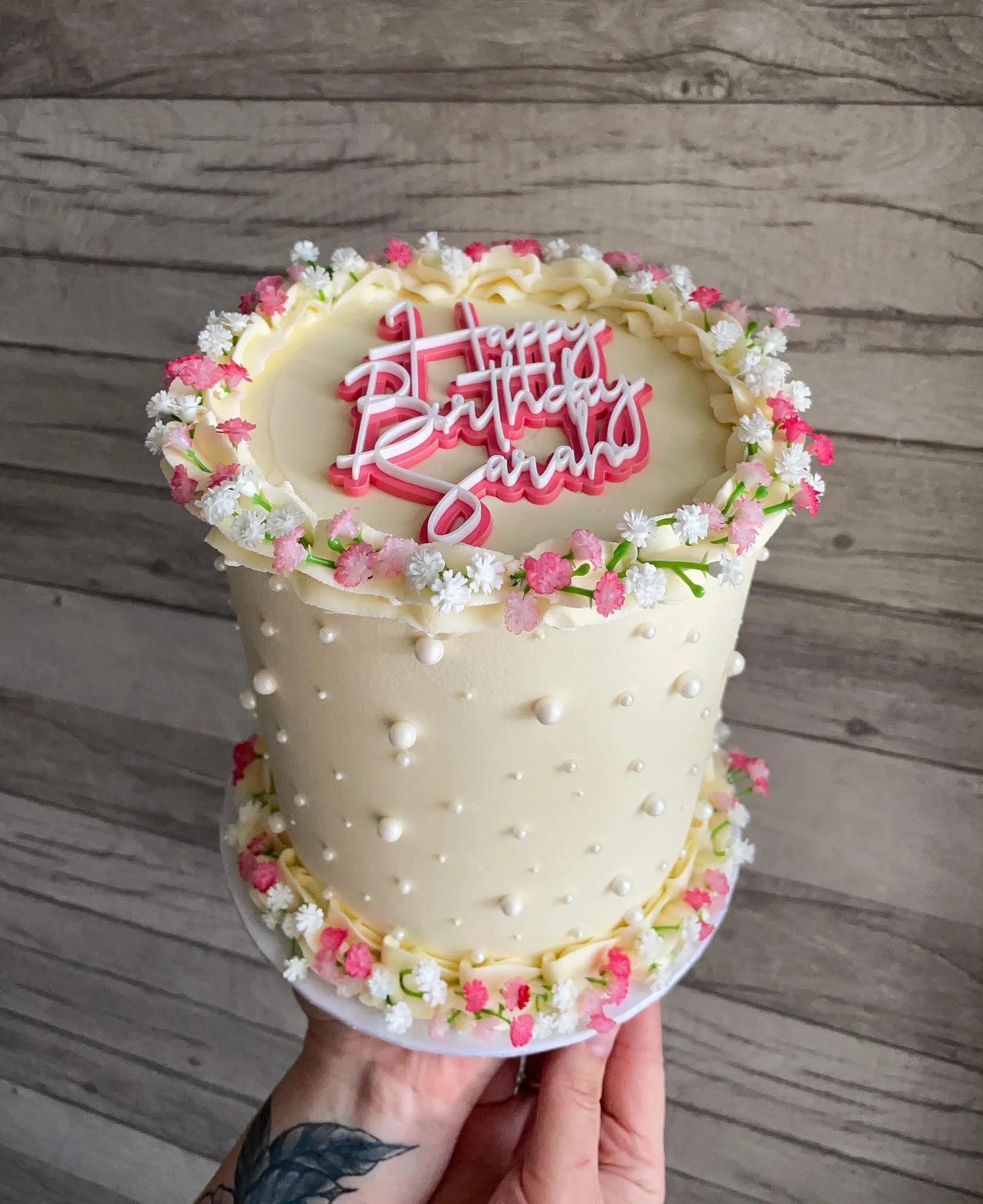 A person holding a birthday cake with white frosting, decorated with small pink and white flowers, and a pink and white "Happy Birthday Sarah" cake topper. The background is wooden flooring.