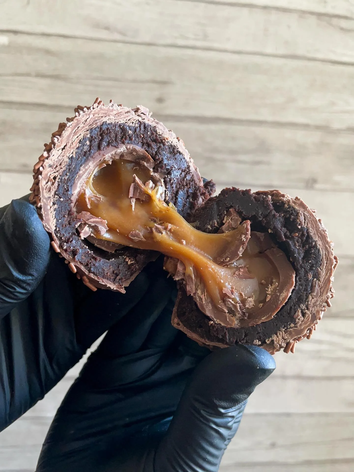 A hand wearing a black glove holding a chocolate and caramel candy with a soft caramel filling.