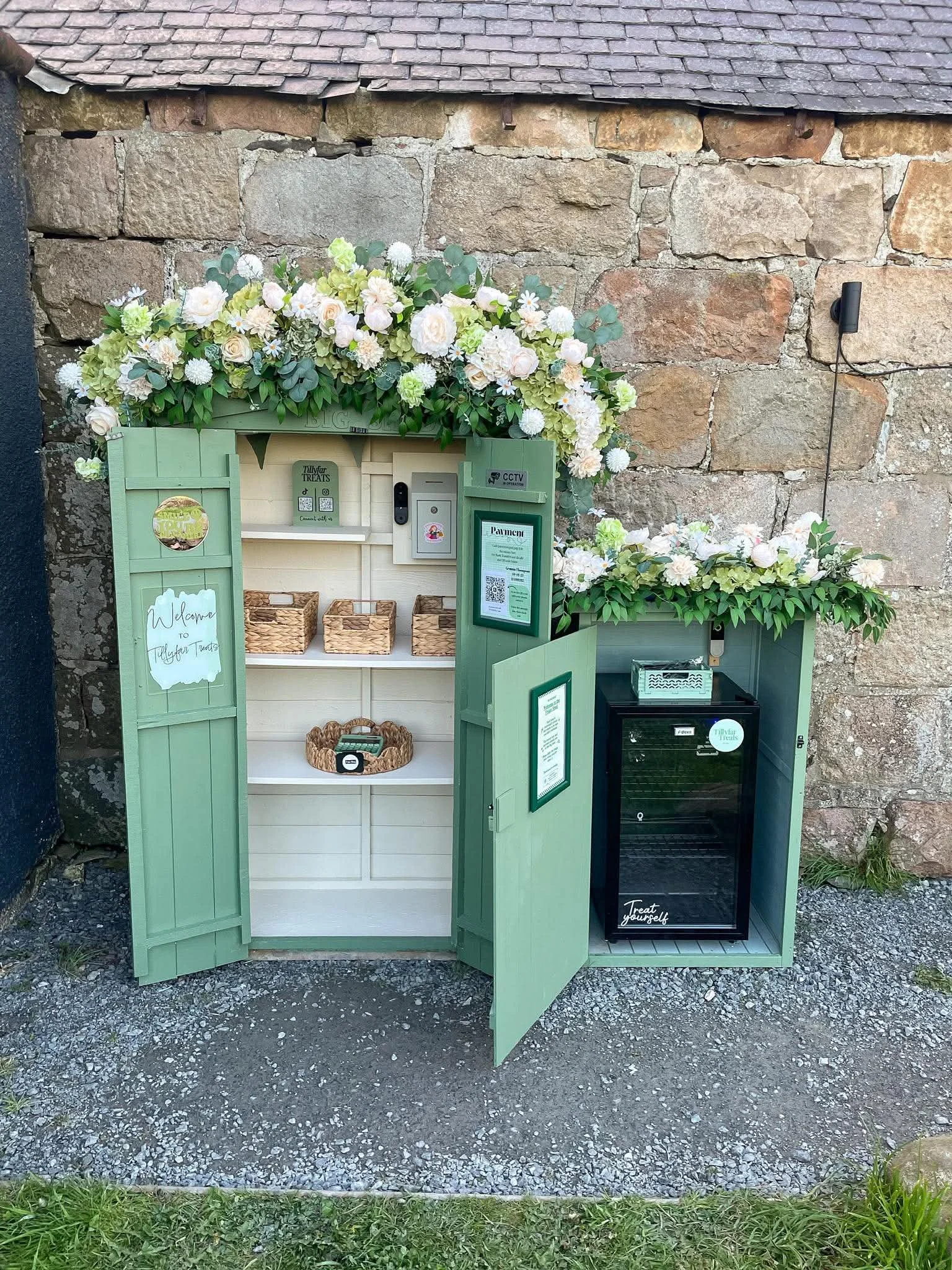 A small green wooden display cabinet with open doors, decorated with white and green flowers on top. Inside the cabinet, there are small baskets, a sign that reads 'Welcome to Tillyfr Trea', QR codes, and a small electronic device. To the right, there is a black mini fridge with a label 'Treat yourself' inside a brick-walled outdoor setting.