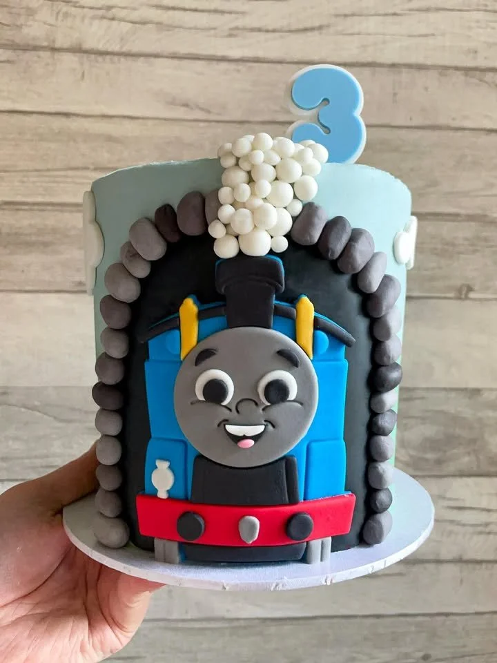 A birthday cake decorated with a Thomas the Tank Engine design, with a blue number 3 candle and white candies on top.