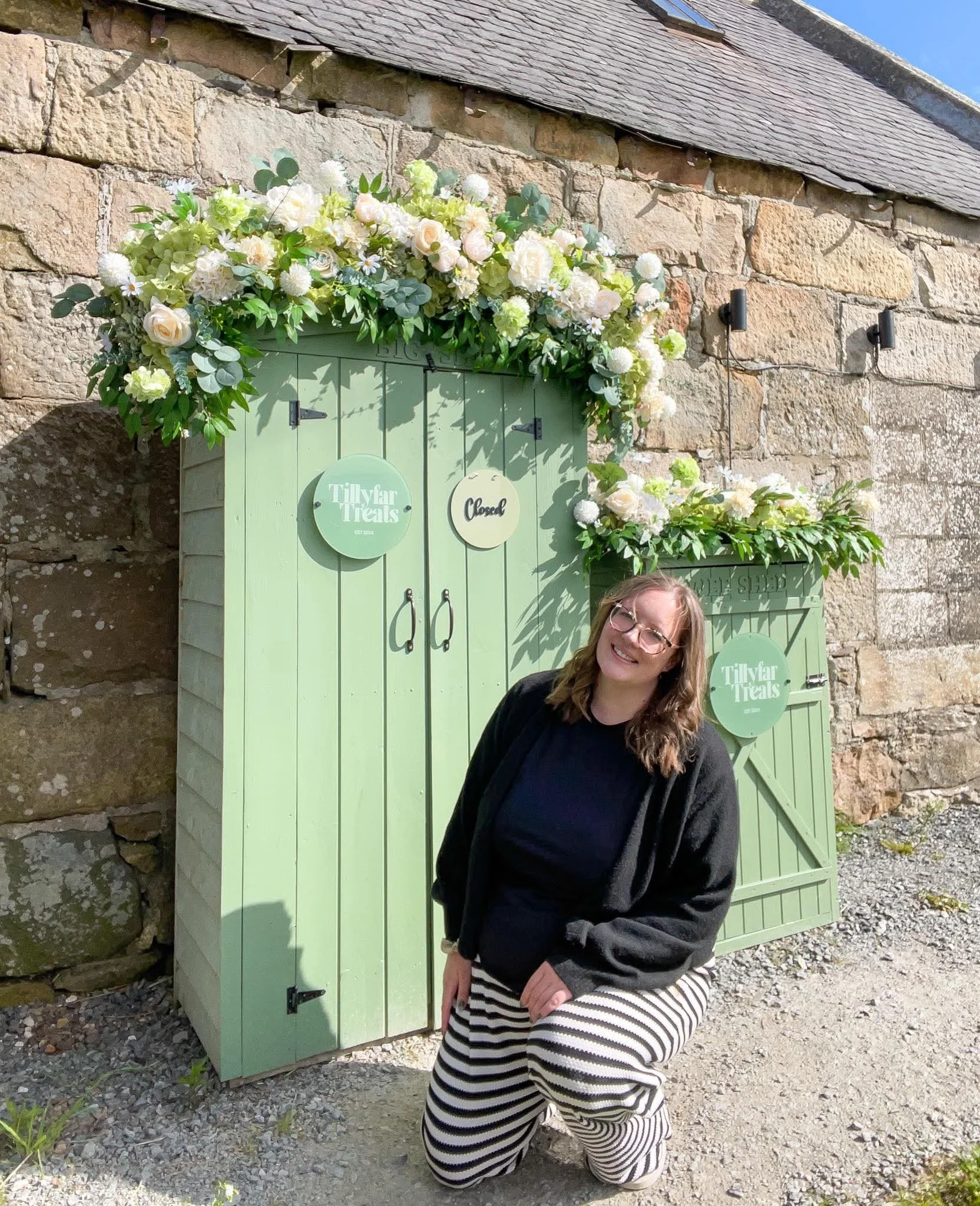 A woman with glasses and long hair smiling, kneeling in front of a green wooden structure decorated with white and light-colored flowers arranged as a floral display, against a stone wall background.
