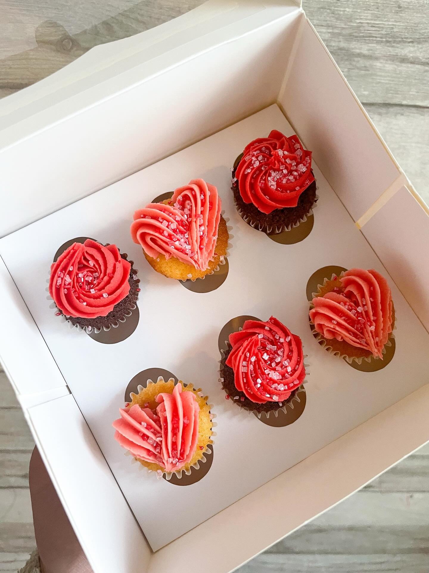 A white box of assorted cupcakes with pink and red frosting and sugar sprinkles.