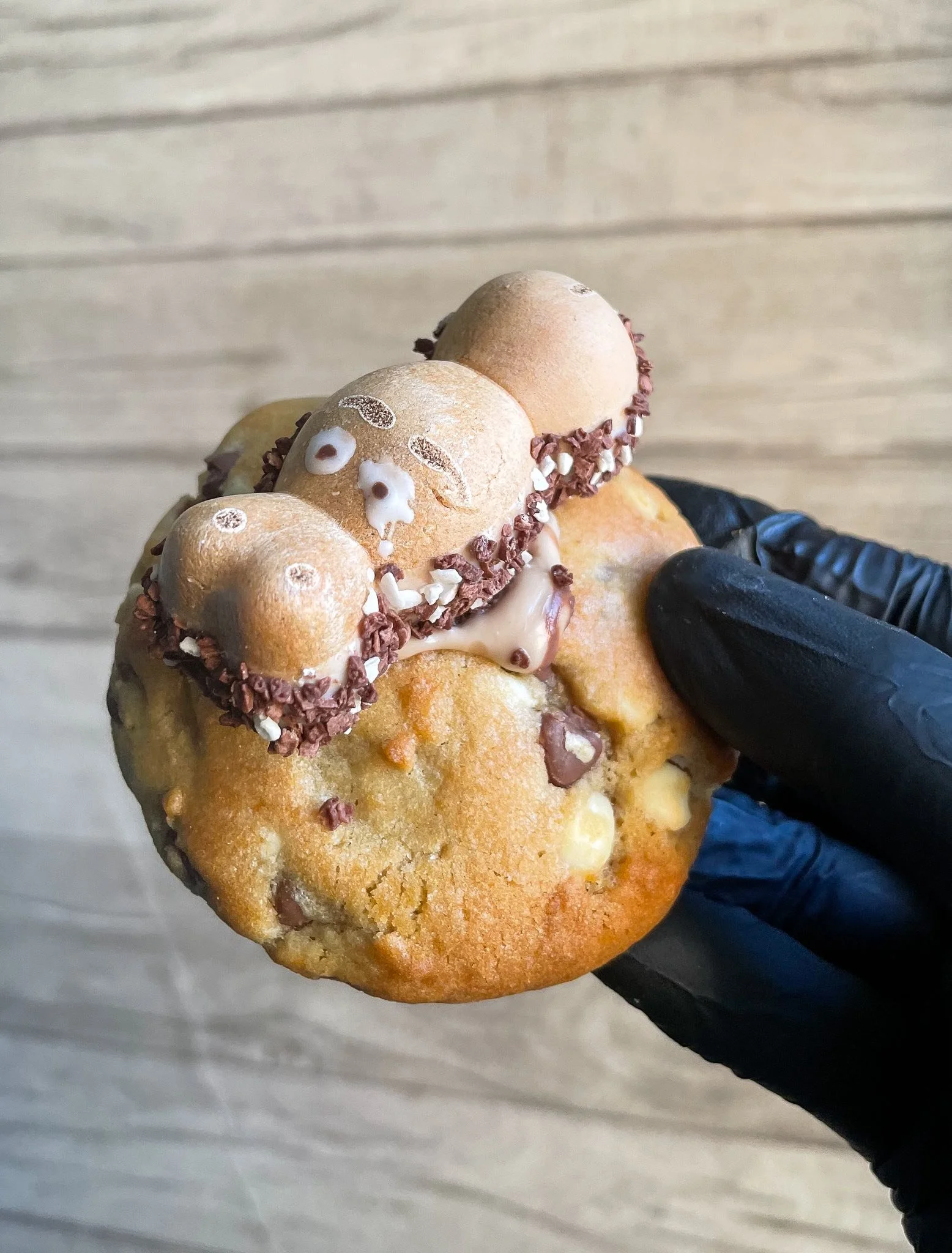 A gloved hand holding a pastry topped with chocolate sprinkles and white icing decorations, set against a light wooden surface background.