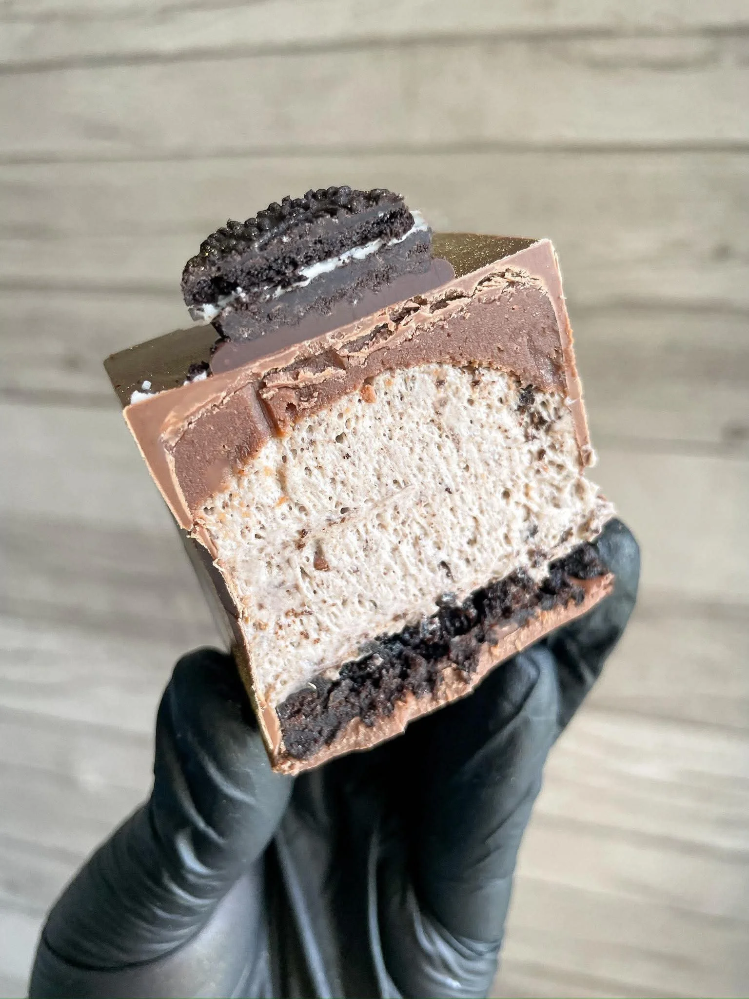 Close-up of a sliced layered dessert with cream and chocolate, topped with a mini Oreo cookie, held by a person wearing black gloves.