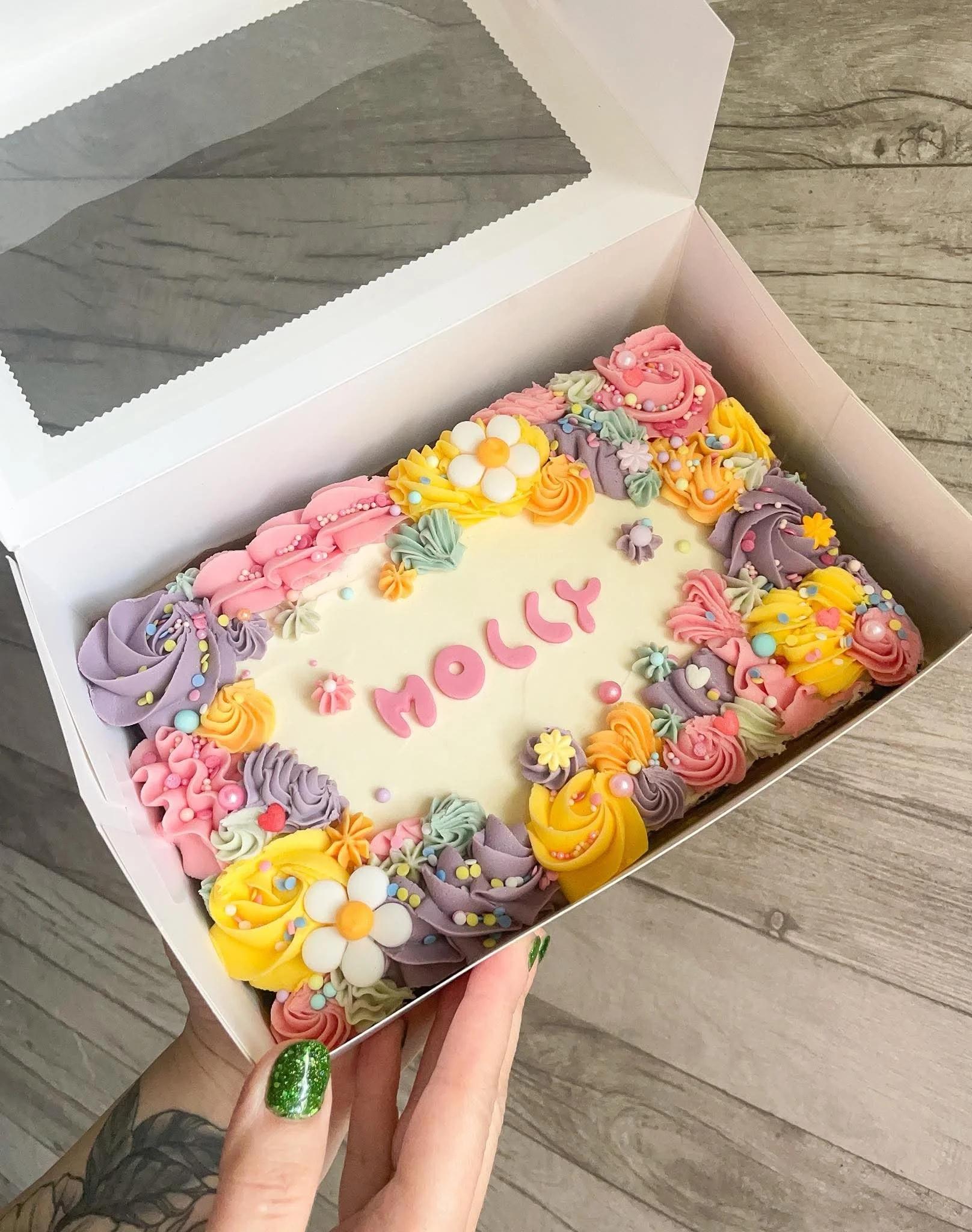 A person's hand with a green glittery nail polish holding a decorated birthday cake inside a white box. The cake has colorful frosting, flowers, and sprinkles on top.