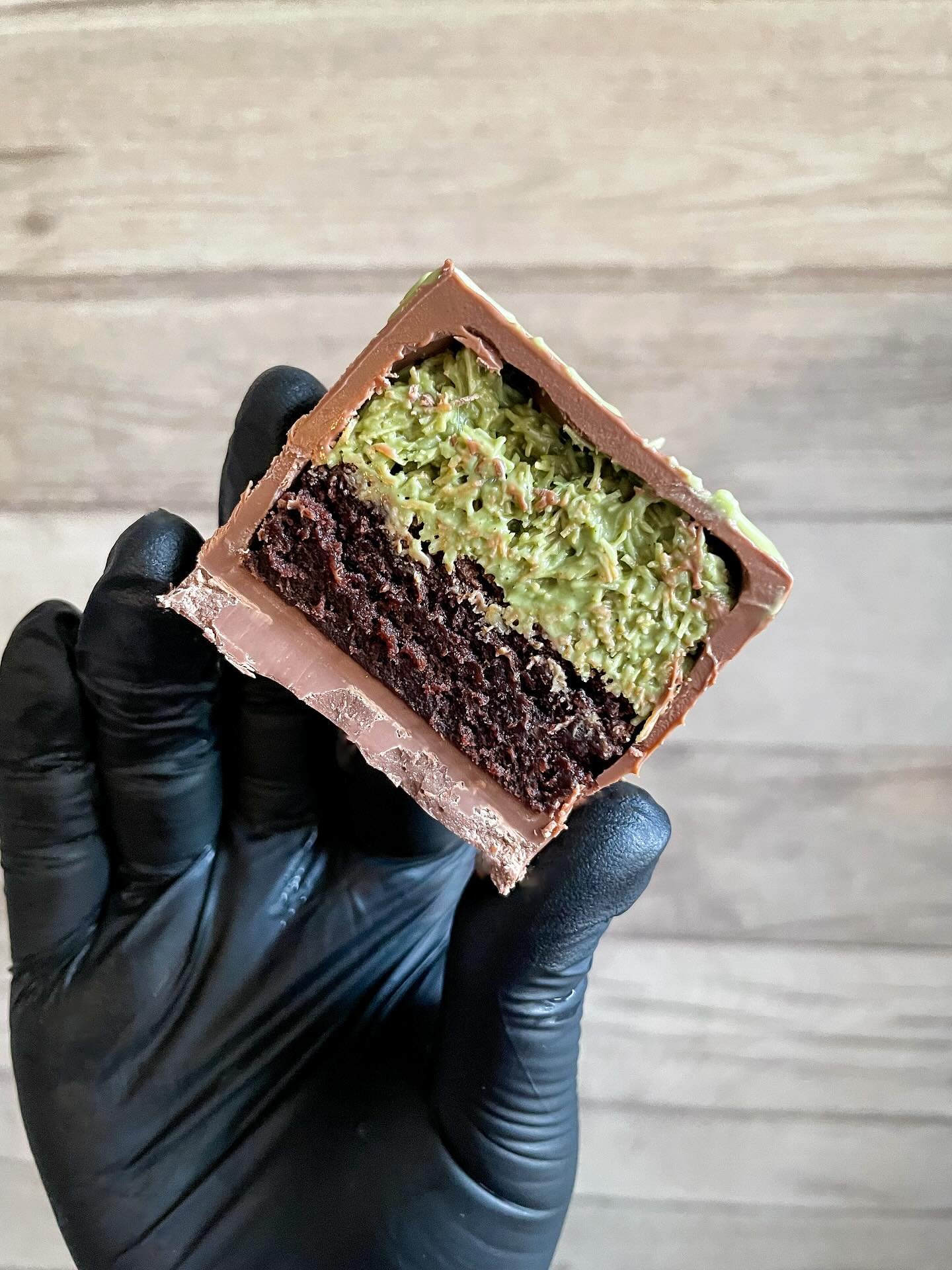 Close-up of a person wearing black gloves holding a layered chocolate and green-colored cake slice on a wooden background.