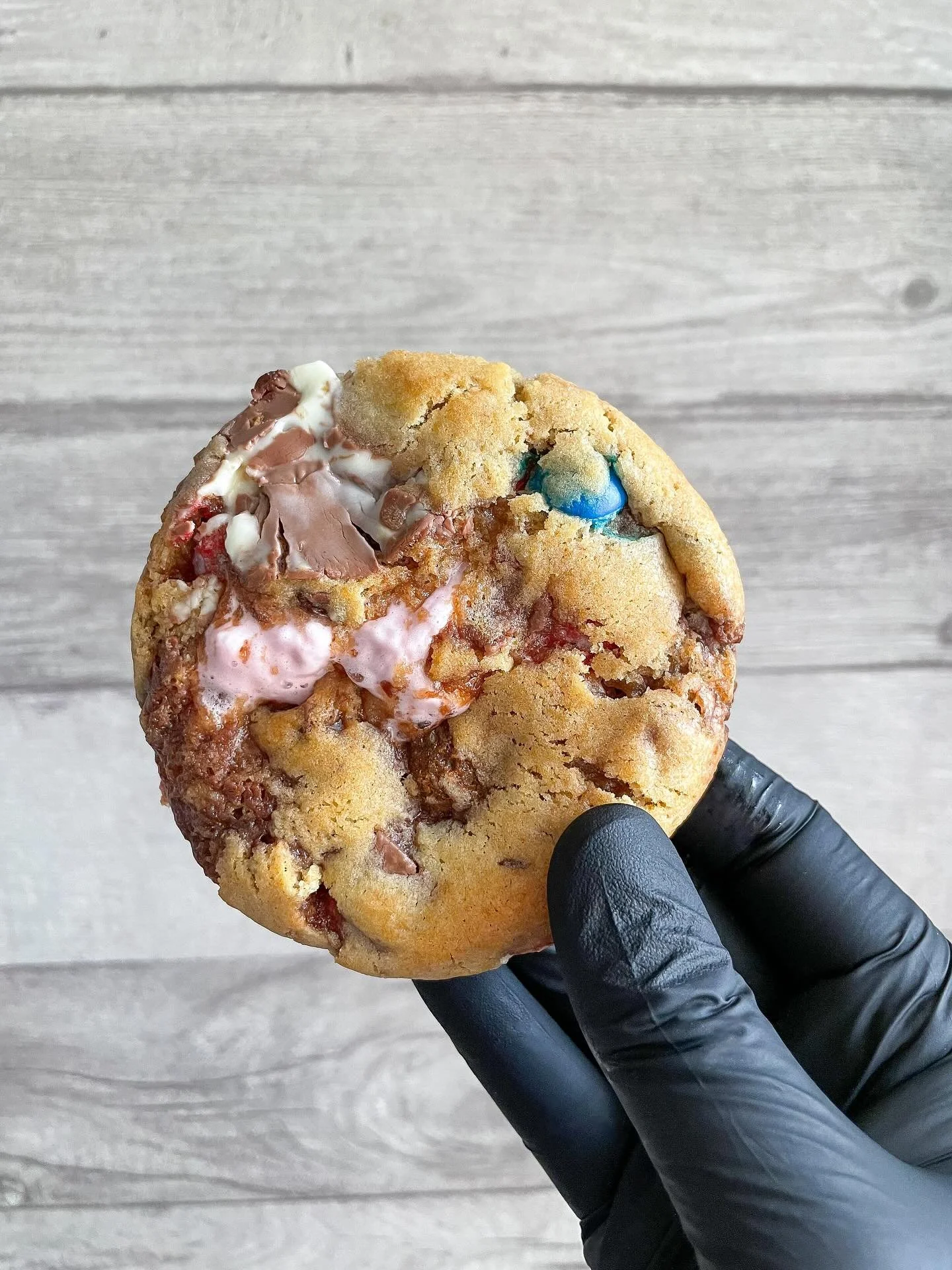 Close-up of a partially eaten cookie with chocolate, marshmallow, and a blue candy on a black gloved hand against a light wooden background.
