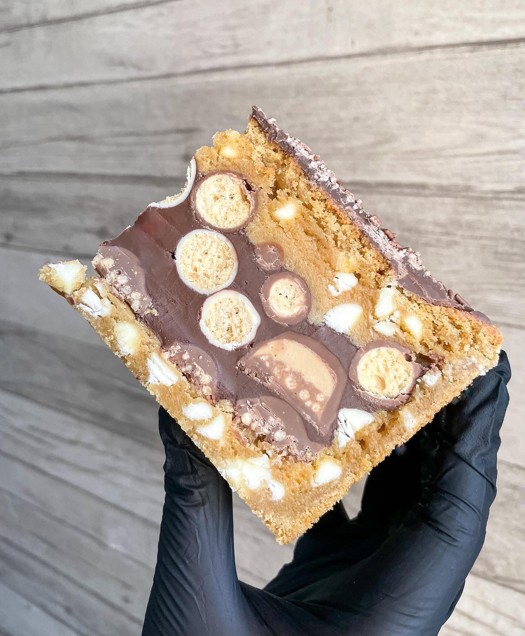 A person wearing black gloves holding a slice of cookie and cream ice cream bar with cookie pieces and white cream filling, against a gray wooden background.